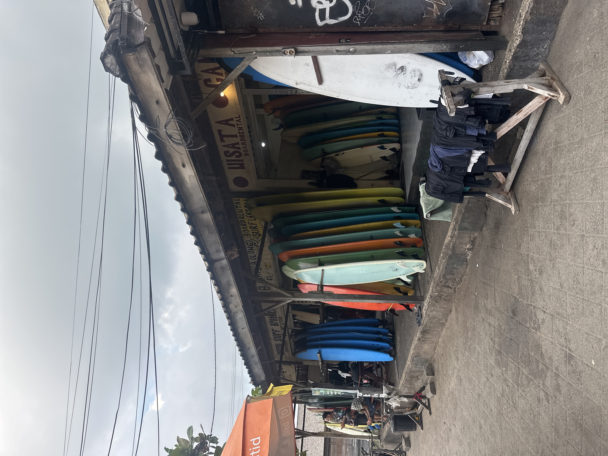 In North Kuta, Indonesia, a beachfront surf shop displays a vibrant collection of surfboards for rent and lessons. An orange awning covers the entrance, and signs advertise "WISATA" and "BOARDRENTAL" in English. A variety of surfboards, in different colors and sizes, are neatly arranged both inside and outside the shop. A wooden drying rack in front of the shop holds several t-shirts and towels. Two men are visible near the entrance of the shop, one is standing and looking at his phone, while the other is sitting down. The ground in front of the shop is paved with concrete tiles, and power lines crisscross the cloudy sky above. The overall scene suggests a bustling tourist area with a focus on water sports.