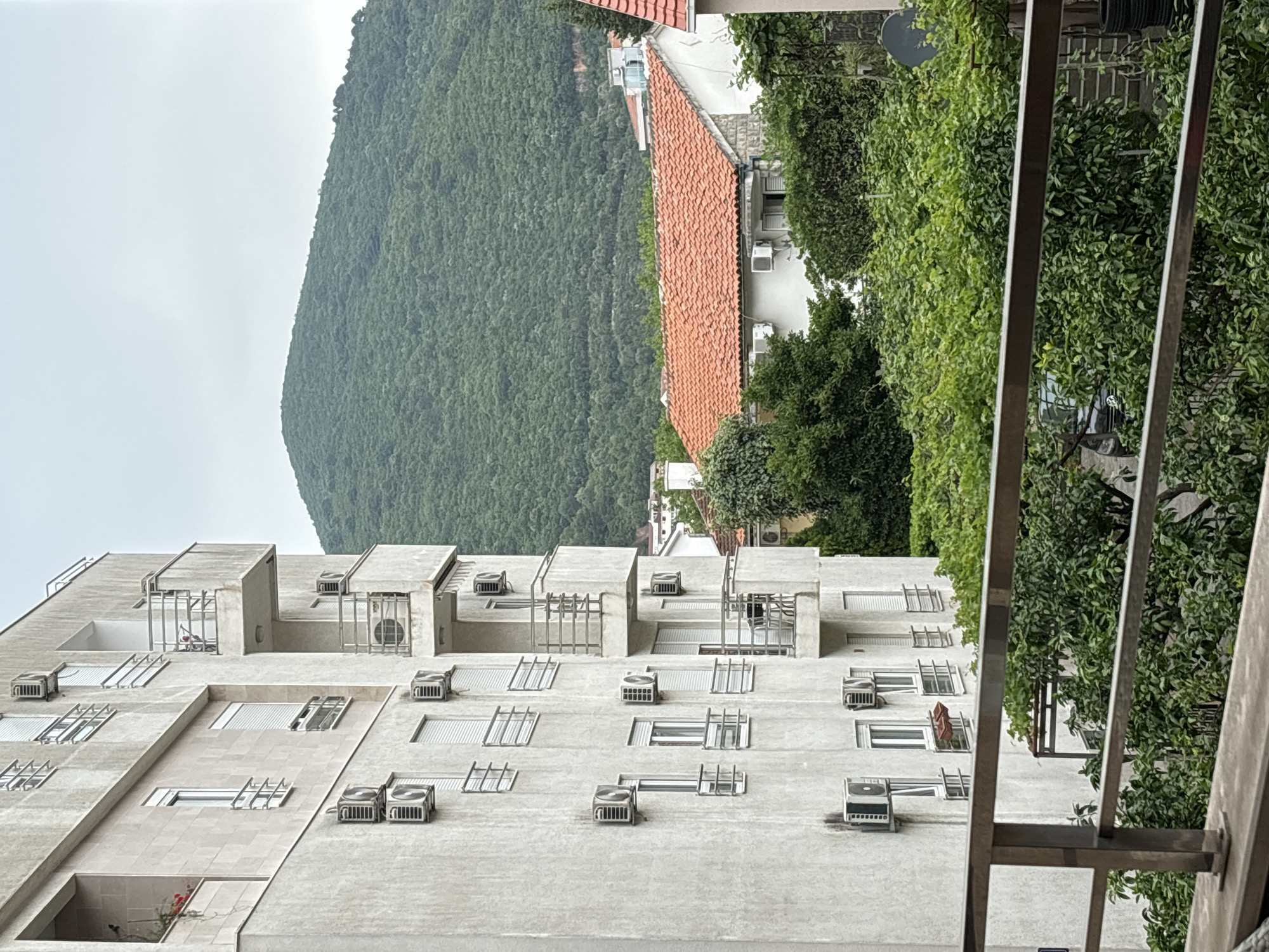 The image captures a detailed urban landscape in Budva, Montenegro, viewed from a balcony or elevated position. In the immediate foreground, a metallic railing runs along the bottom, framing the scene.
The central subject is a modern, multi-story building constructed from light-grey concrete. Its facade is characterized by numerous rectangular windows, some featuring metal grilles, and a multitude of external air conditioning units attached to its walls. The building exhibits a stepped architectural design with various recessed and protruding sections. Small red flowers can be seen in a planter on one of its lower levels, adding a touch of color to the otherwise monochromatic exterior.
To the right of the concrete building, a dense cluster of lush green trees fills a significant portion of the midground. Nestled amongst this foliage, parts of a smaller, traditional building with a distinctive orange-tiled roof are visible. A small, dark satellite dish can be spotted above the treetops further to the right.
Dominating the background on the left is a large, heavily forested mountain, its slopes covered in dense green vegetation. The sky above is uniformly light grey and overcast, suggesting a cloudy day with diffused light. No specific activity or people are visible in the static scene. There is no discernible text within the image.