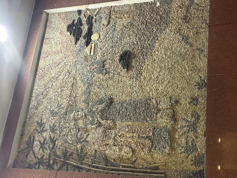 This image displays a large, rectangular pebble mosaic art piece set into a wall in Ngũ Hành Sơn Ward, Vietnam. The mural depicts a traditional Vietnamese landscape scene. On the left, stylized dark bamboo stalks with leaves extend upwards. In the center, two human figures, rendered in traditional attire using varying shades of pebbles, stand facing each other, suggesting an interaction or conversation. The figure on the left is adorned in lighter, brownish-red hues, while the figure on the right is in darker, gray-brown tones. To the right of these figures, a smaller individual rides an animal, possibly an ox or water buffalo, depicted in dark pebbles. Further right and slightly lower, two stylized houses or huts with distinctive roofs are visible. The upper right portion of the mural illustrates a sky or sunrise/sunset with radiating lines created by alternating light and dark pebbles. The entire landscape, including the ground and undulating hills, is composed of numerous small, rounded pebbles of cream, light brown, and gray hues. The mosaic is framed by dark reddish-brown polished material at the top, bottom, and right, with an overhead light source visible in the upper right corner of the overall frame.