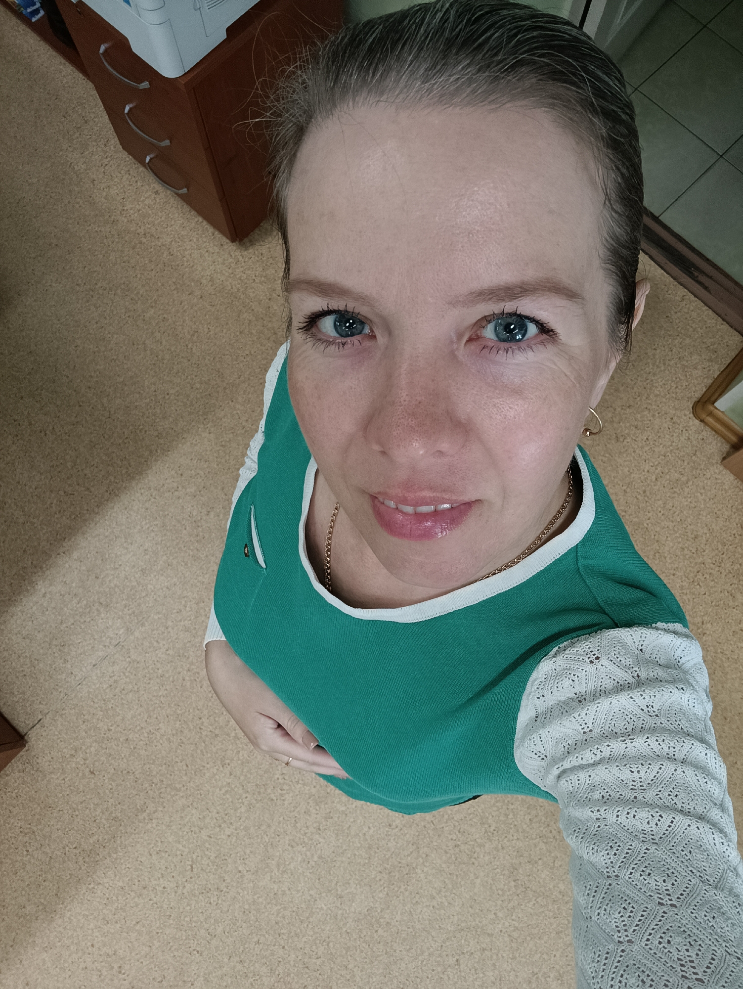 A woman is pictured in a top-down, high-angle close-up shot within an interior setting. The woman has fair skin, blue eyes, and her hair is pulled back. She is wearing a green top with white trim and sleeves with a lace-like texture. A gold chain is visible around her neck, and she wears small hoop earrings. The background includes a beige patterned floor, a brown cabinet with drawer pulls, and a section of a doorway. The location is possibly in an office or domestic environment within the city of Chaikovsky, Russia.
