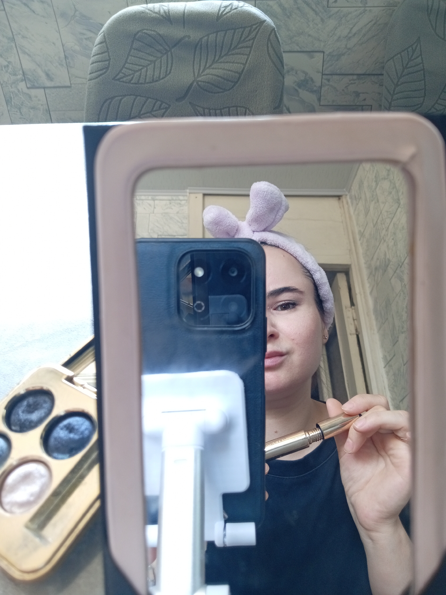 A woman with a purple headband on her head is taking a selfie in a mirror. She is holding a gold mascara wand in her right hand and a phone in her left hand. In the foreground, a gold eyeshadow palette with four circular eyeshadows is visible. The background shows a tiled wall and a patterned chair. The scene appears to be in a bathroom or dressing area. The image is taken in Khujand, Tajikistan.