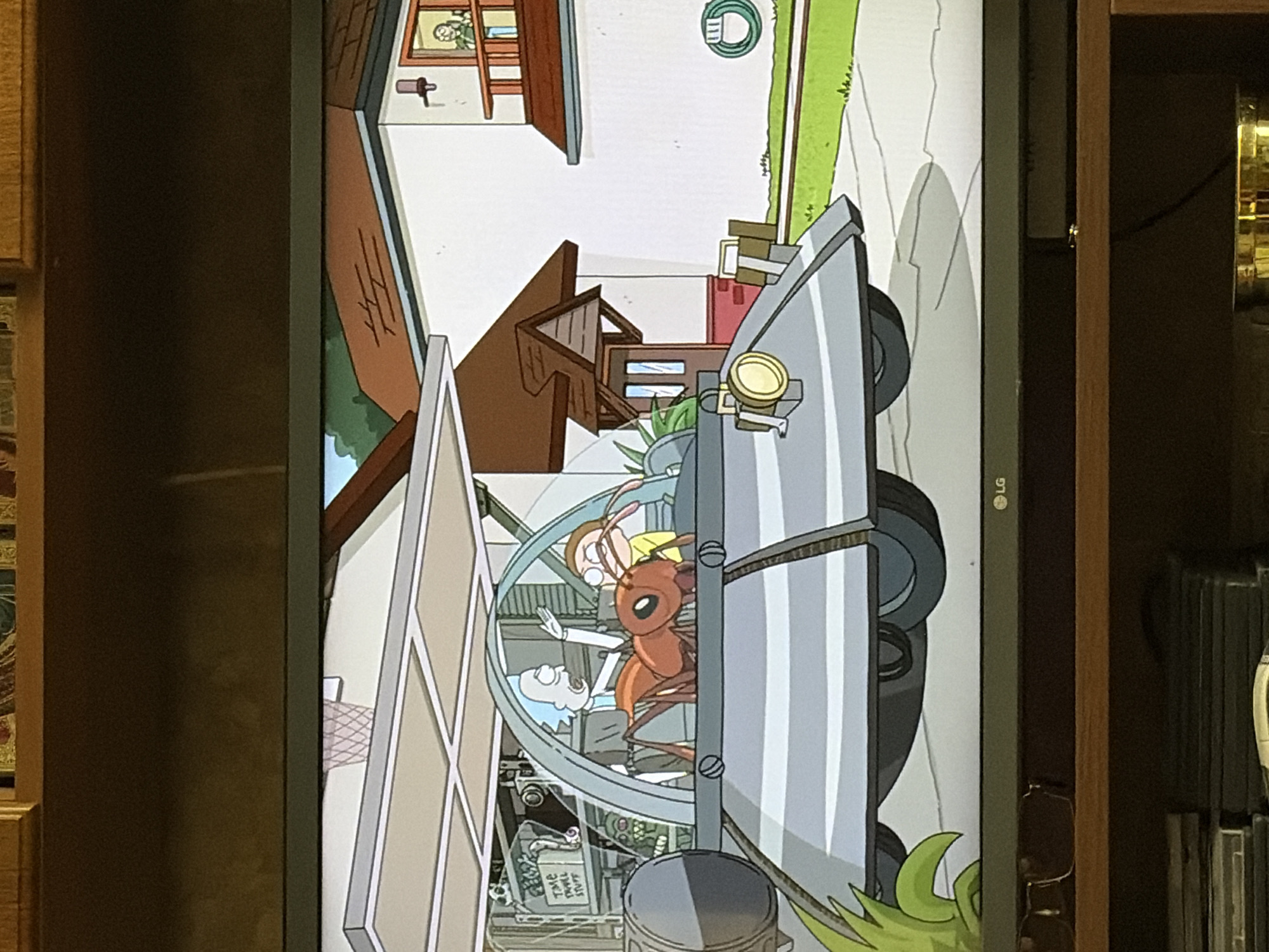 In an animated scene set in the city of Unknown, Unknown, Rick Sanchez and Morty Smith are inside a large, ant-shaped vehicle. Rick, with his signature blue hair, is in the driver's seat, gesturing with his right hand. Morty, wearing a yellow shirt, sits beside him. The vehicle's interior appears to be a laboratory, with shelves visible behind Rick containing equipment and a sign that reads "Time Travel Stuff." Outside the vehicle, a suburban house is visible, with a person looking out of an upstairs window. The vehicle is parked on a driveway in front of the house.