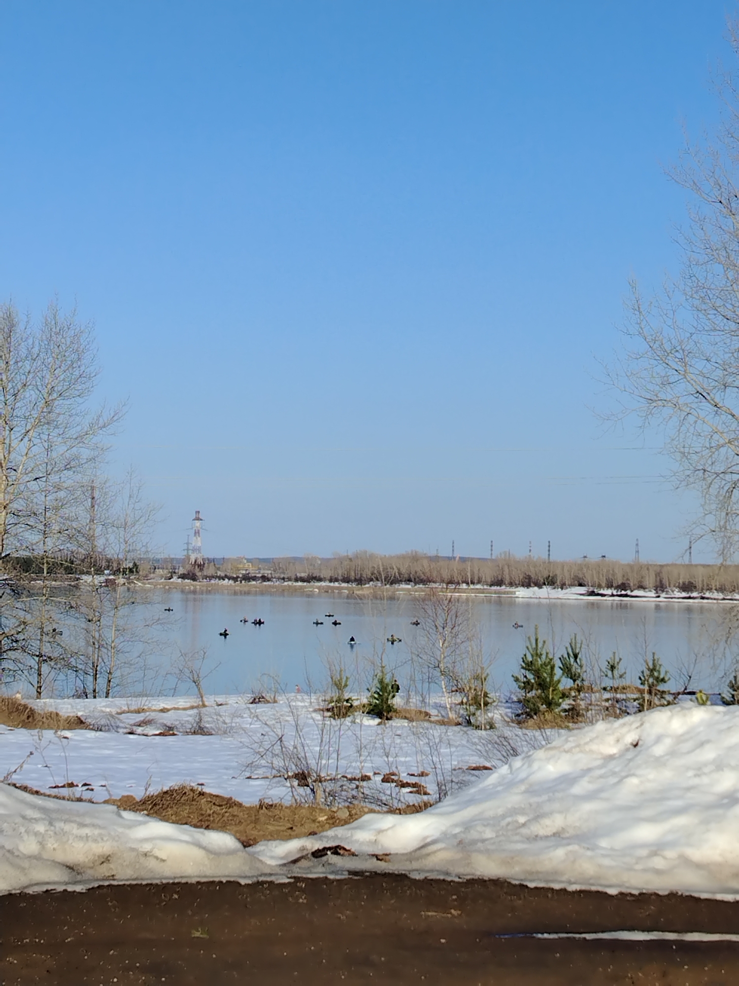 The image presents a landscape in a location identified as Noviy, Russia. It is a daytime scene under a clear, blue sky. The foreground features a dark, possibly wet or muddy surface, with a snowdrift separating it from the middle ground.
The middle ground consists of a body of water, likely a river or lake, with reflections of the sky and distant trees. Several small watercraft, possibly kayaks or canoes, are visible on the water. The shoreline is marked by patches of snow and small evergreen trees.
In the background, a line of bare trees is visible along the opposite shore. Power transmission towers and other industrial structures are present on the horizon. Two bare trees are also present at the left and right edges of the image.