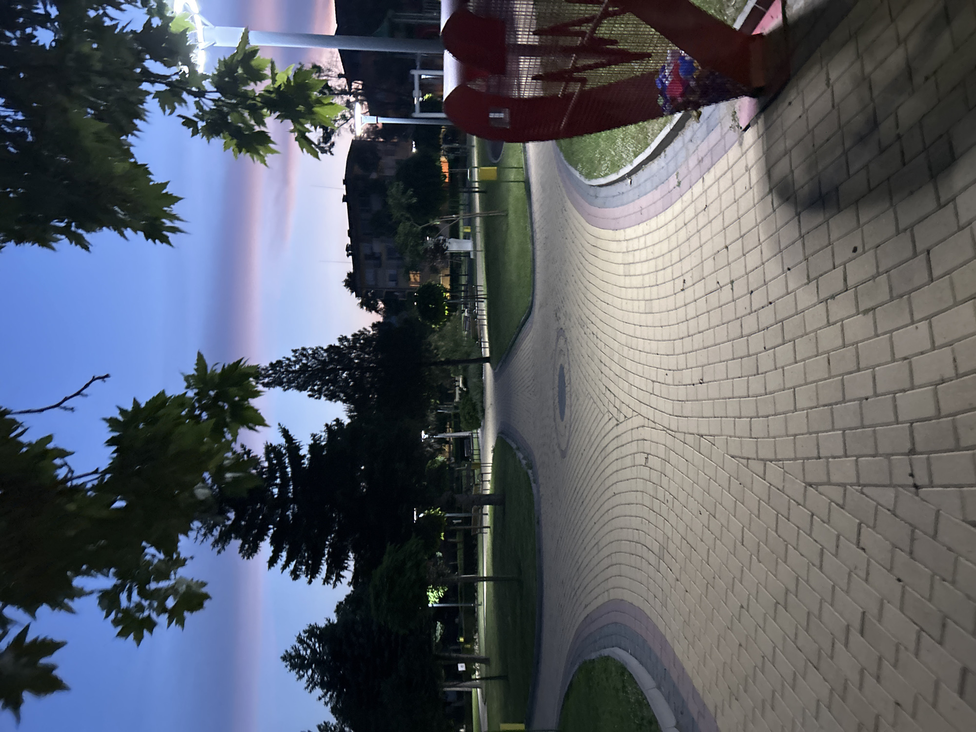 The image depicts a park in Dzhebel, Bulgaria, at twilight, characterized by a sky transitioning from deep blue overhead to soft pink and purple hues near the horizon. The park features a network of paved pathways constructed from light-colored interlocking pavers, with darker decorative strips along the edges, forming curves and intersections. Lush green lawns border these pathways, which are lined with various trees, including dark evergreen conifers and deciduous trees. Multiple modern park lights with white LED illumination are active, casting light onto the paths and surrounding vegetation, creating distinct shadows.
In the immediate foreground on the right, a large, red, metallic, heart-shaped structure is present. This structure features cutout letters, appearing to be a stylized "WM" or similar design, and functions as a collection bin, containing colorful plastic items visible in its lower section. Further into the park, several benches are visible along the pathways and on the grass. A yellow cylindrical trash receptacle is situated near a path intersection in the midground. In the more distant background, beyond the park's trees, dark silhouettes of multi-story buildings are visible against the twilight sky. No individuals are present in the scene.