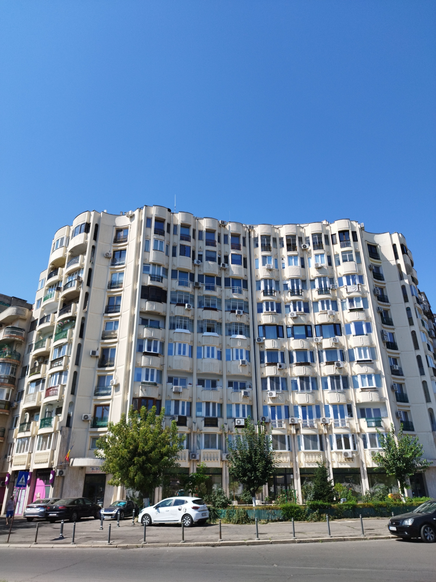 An exterior, eye-level, full shot captures a large, cream-colored, curved apartment building under a clear blue sky in Bucharest, Romania. The building features numerous balconies and windows, with several air conditioning units visible on its facade. The ground level of the building appears to house businesses, with large glass windows and awnings. Trees and landscaping are situated along the sidewalk in front of the building. Several cars are parked on the street and in front of the building, including a white sedan, a black sedan, and a silver sedan. A pedestrian crossing sign and a few street poles are also visible on the sidewalk. The overall scene suggests a sunny day in an urban environment.