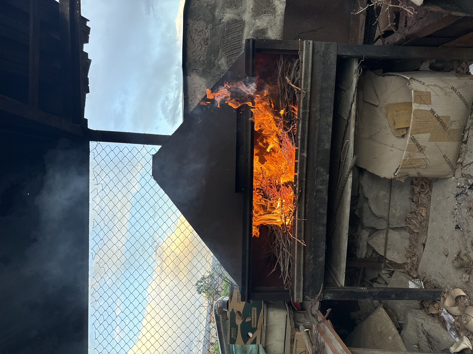 A fire burns inside a black metal furnace. Small branches are burning. Smoke is visible. Adjacent to the furnace is a partially crushed cardboard box with "WILDBERRIES" printed on its side. A chain-link fence runs along the left side of the frame, behind which is a view of a cloudy sky. The location is Yerevan, Armenia.