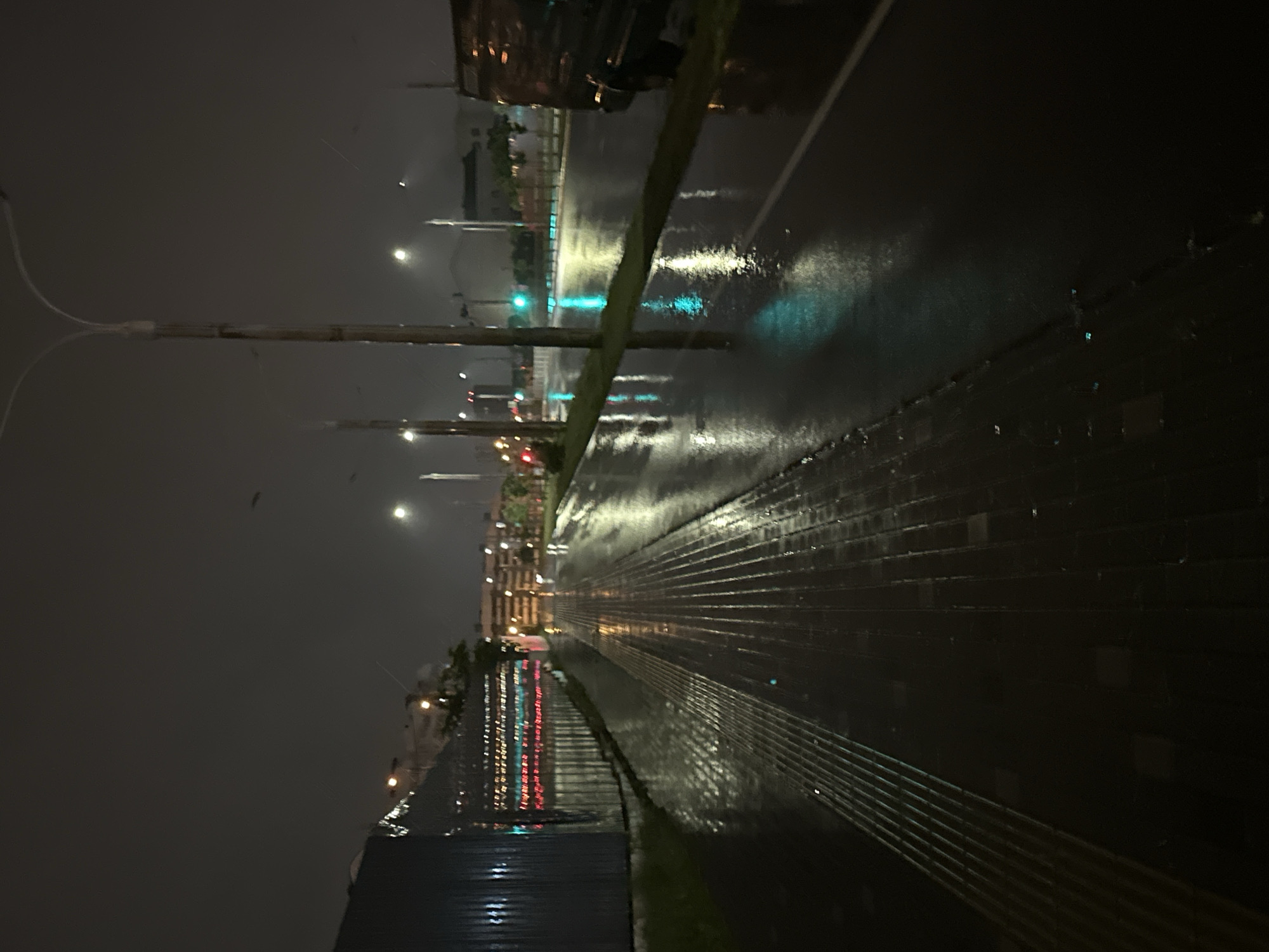 The image captures a rain-slicked street at night, likely in or near Бараўлянскі сельскі Савет, Belarus. The scene is dominated by reflections on the wet asphalt and a paved walkway, illuminated by streetlights and distant building lights. The walkway, to the left of the frame, is made of patterned bricks or pavers and shows parallel raised lines. To the right, a dark, possibly a van or truck, is partially visible. The road features a white lane marker. In the background, several streetlights are visible, with one emitting a bright white light. Further back, the lights of buildings are scattered across the dark sky. Some red and green lights are also visible in the distance, possibly from traffic signals or signage. The overall atmosphere is dark and reflective due to the nighttime setting and the recent rainfall.