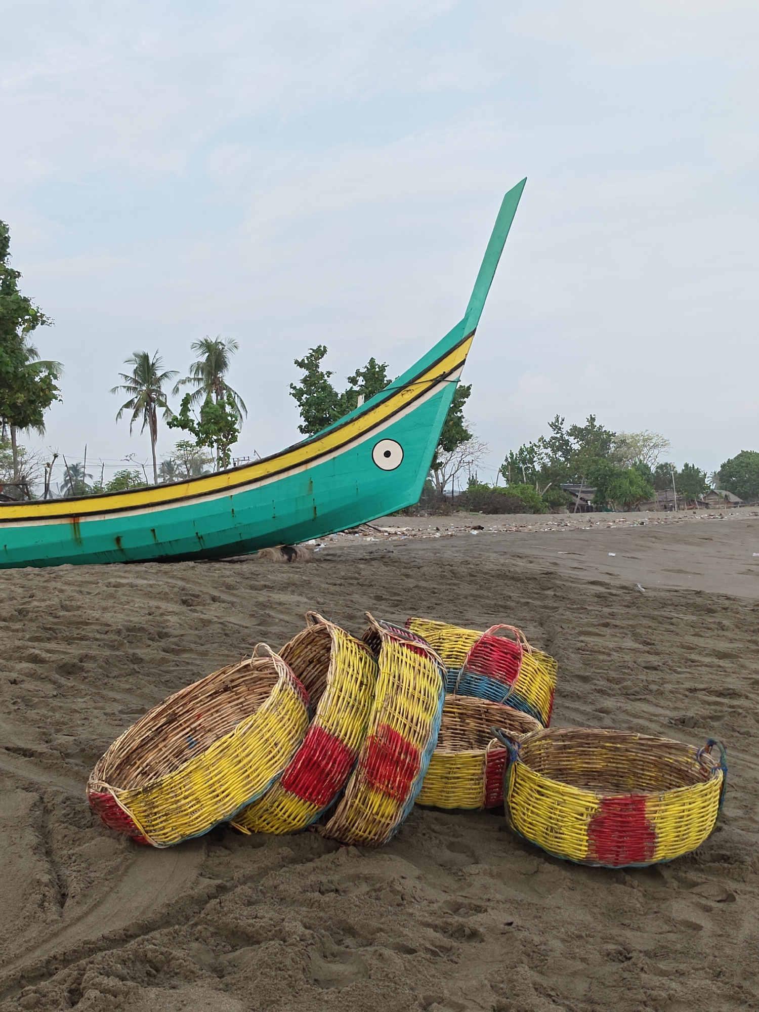 The image depicts a beach scene with a traditional fishing boat and several woven baskets on the sand. The boat, predominantly turquoise with yellow and red accents, is partially visible on the left side, its distinctive pointed bow facing upwards. In the foreground, a cluster of woven baskets, some decorated with vibrant yellow and red patterns, are arranged on the sandy beach. In the background, palm trees and other vegetation dot the shoreline, with a few scattered structures suggesting a settlement. The sky above is overcast, with a light cloud cover. The scene appears to be located in Curehtunong, Indonesia, based on the provided context. There are no people or animals visible in the image.