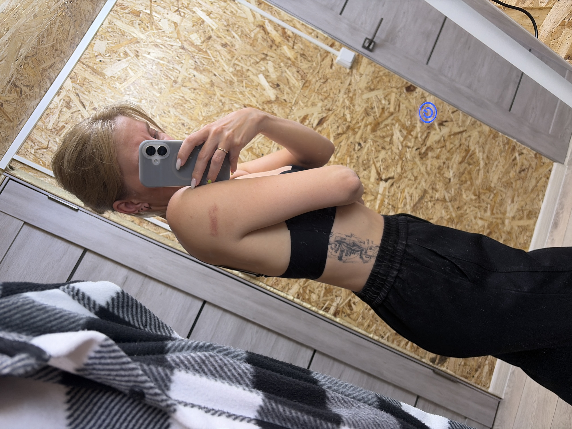 A person is taking a selfie in what appears to be a fitting room or bedroom. The individual, with blonde hair, is holding a grey smartphone and wearing a black sports bra and black pants. The setting suggests a home or temporary lodging, characterized by a wooden wall with visible patterns. The foreground includes a blurred black and white checkered fabric, probably a blanket or part of the bedding. Location information (Chaikovsky, Russia) is unavailable from the visual content alone.