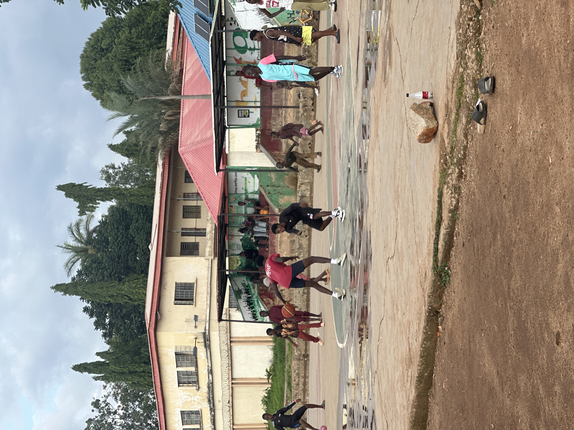 This is a medium shot of a group of young men playing basketball on an outdoor court in Jos, Nigeria. The court is wet and has puddles from recent rain. The players are wearing various athletic attire, including t-shirts, shorts, and sneakers. One player in a red shirt is dribbling a basketball, while others are positioned around the court. In the background, there are buildings with red and blue roofs, and lush green trees. Spectators can be seen sitting on benches under a shaded structure. The overall atmosphere appears to be casual and recreational.