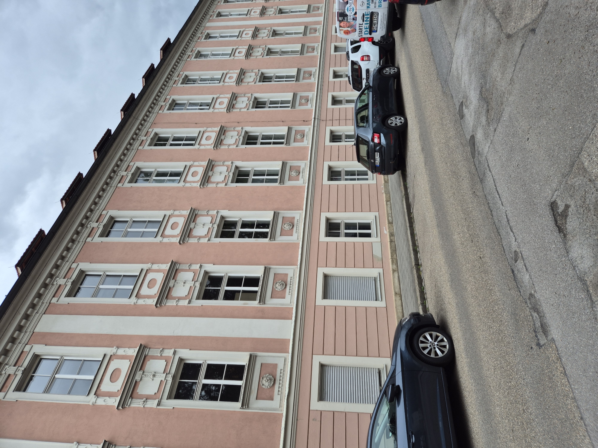 This is a slightly tilted, eye-level shot of a light pink building with white accents, viewed from the street. The building appears to be an apartment complex or office building, with rows of identical windows visible on multiple floors. The windows are mostly white-framed, with some featuring what look like interior blinds. The building's facade is decorated with subtle, ornate details between the windows.
Parked along the street in front of the building are three vehicles: a dark blue hatchback, a white panel van, and another vehicle partially obscured. The panel van has text on its side, including "STARTE DEINE KAF" and "BRUNNER". The license plate on the blue hatchback is visible and reads "OAL TL 444". The street itself is asphalt, with some wear and tear, and a sidewalk is visible along the building's edge.
The sky is overcast, suggesting a cloudy day. The lighting is even, with no strong shadows, consistent with a cloudy sky or possibly late afternoon. The overall impression is of a typical urban scene in Regensburg, Germany, showing the architecture of the building and the presence of vehicles on the street. There are no people clearly visible in the frame.