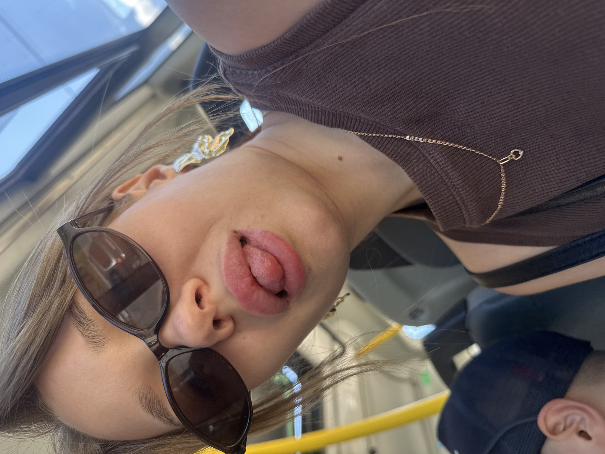 The image is a close-up, low-angle selfie of a young woman with blonde hair and sunglasses, sticking her tongue out. She is wearing a brown ribbed tank top and a gold necklace with a heart pendant. She has large, golden butterfly-shaped earrings. The background appears to be the interior of a public transport vehicle, possibly a bus, with yellow poles and windows showing a bright sky. The woman is in the foreground, with her face tilted towards the camera. A person wearing a dark baseball cap is partially visible in the lower left corner of the frame. The overall lighting is bright, suggesting daytime. The specific location in Chaikovsky, Russia, cannot be determined from the visual information present in the image.