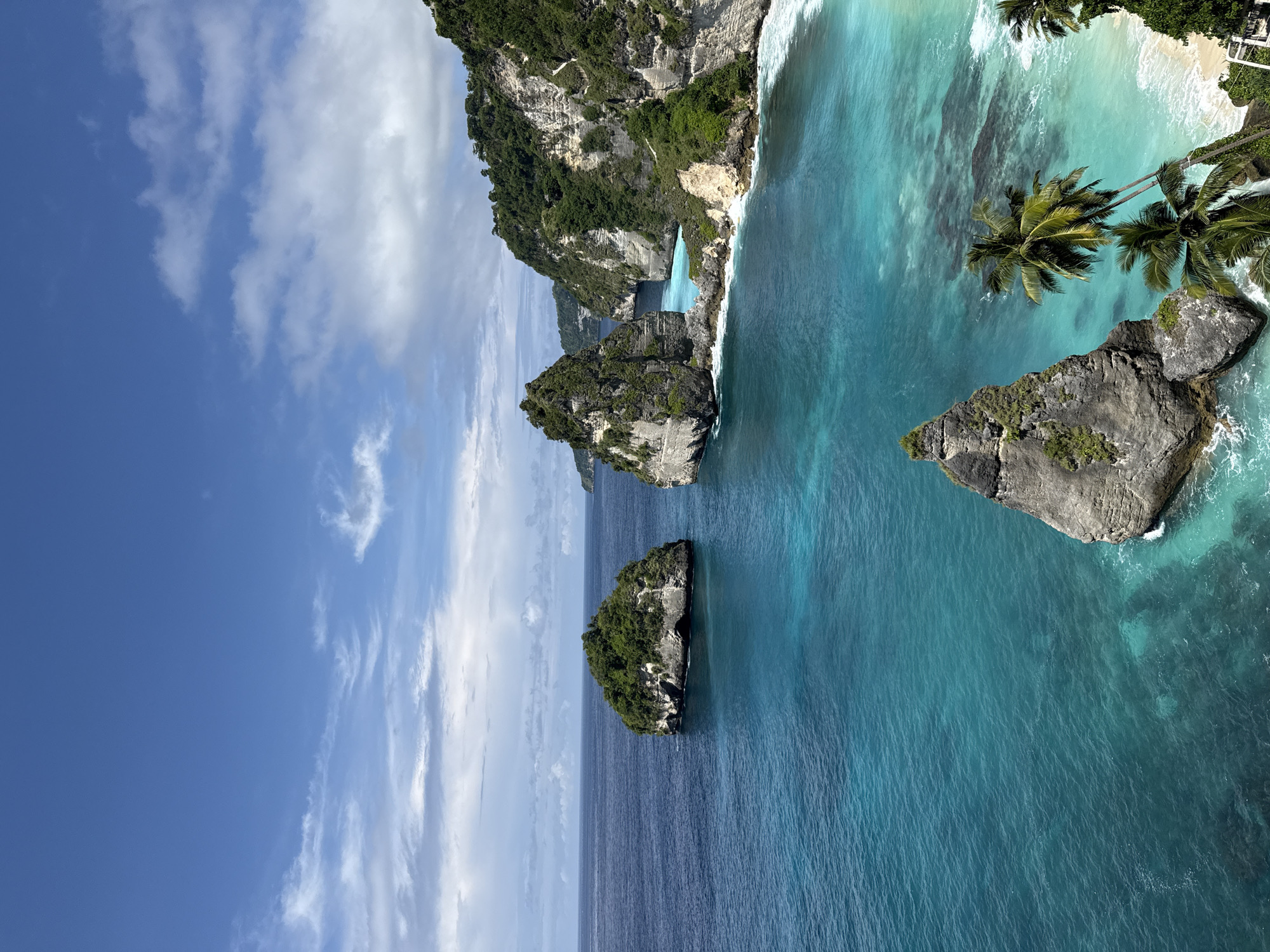 The image captures a coastal scene in Pejukutan, Indonesia, featuring a clear blue sky with scattered white clouds. The turquoise ocean dominates the lower portion, with visible variations in color indicating different depths and wave patterns. Several rocky islands of varying sizes emerge from the sea, covered in green vegetation. Palm trees line the edge of a sandy beach at the top right. The coastline consists of tall cliffs, also covered in dense greenery.