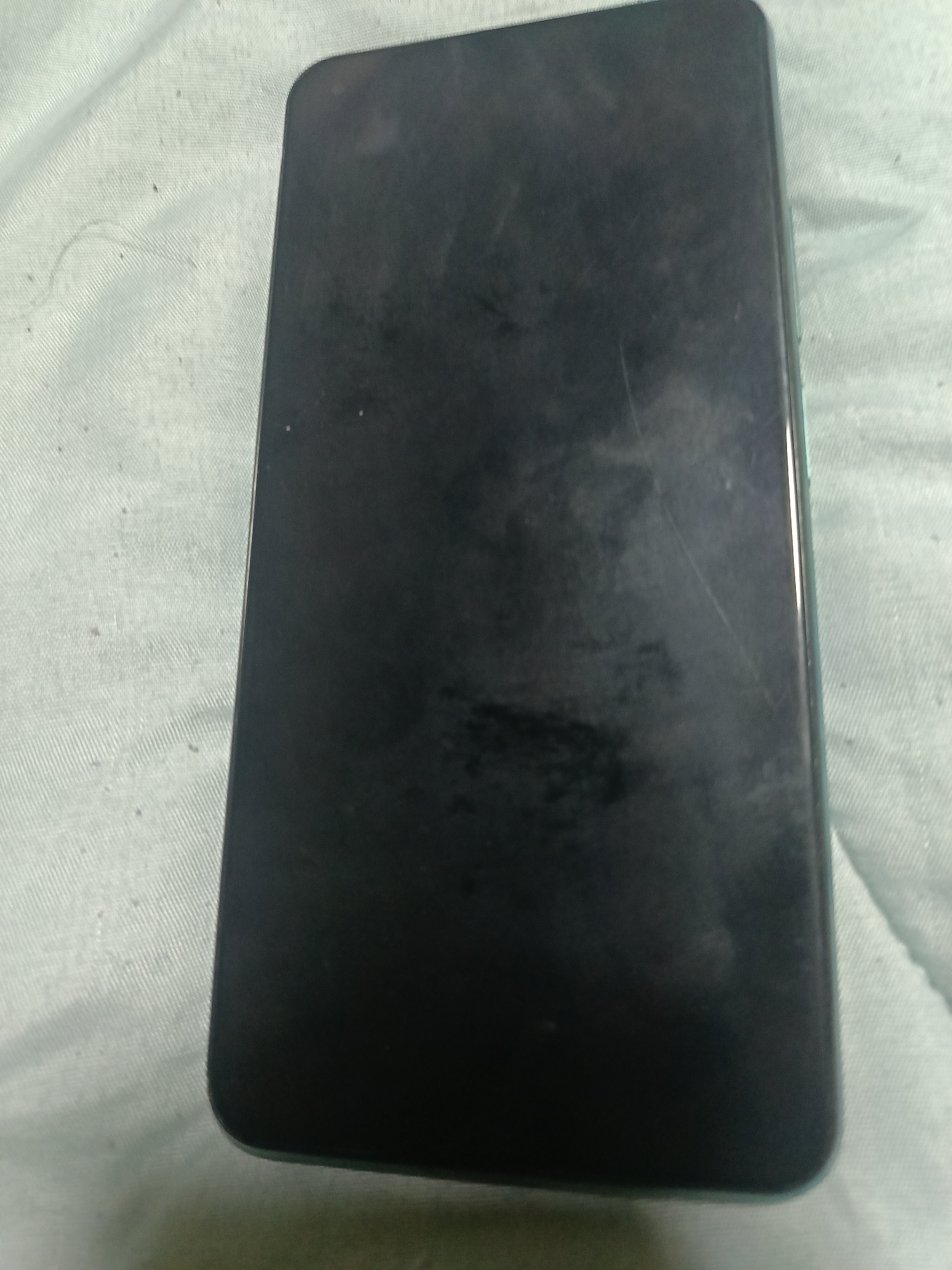 A black smartphone lies on a light blue textured surface, likely fabric, in Mizia, Bulgaria. The phone's screen is facing upwards, showing some smudges and light scratches. The surface beneath the phone appears to be a thin, woven material. There are no people or other objects visible in the image. The lighting suggests an indoor environment.