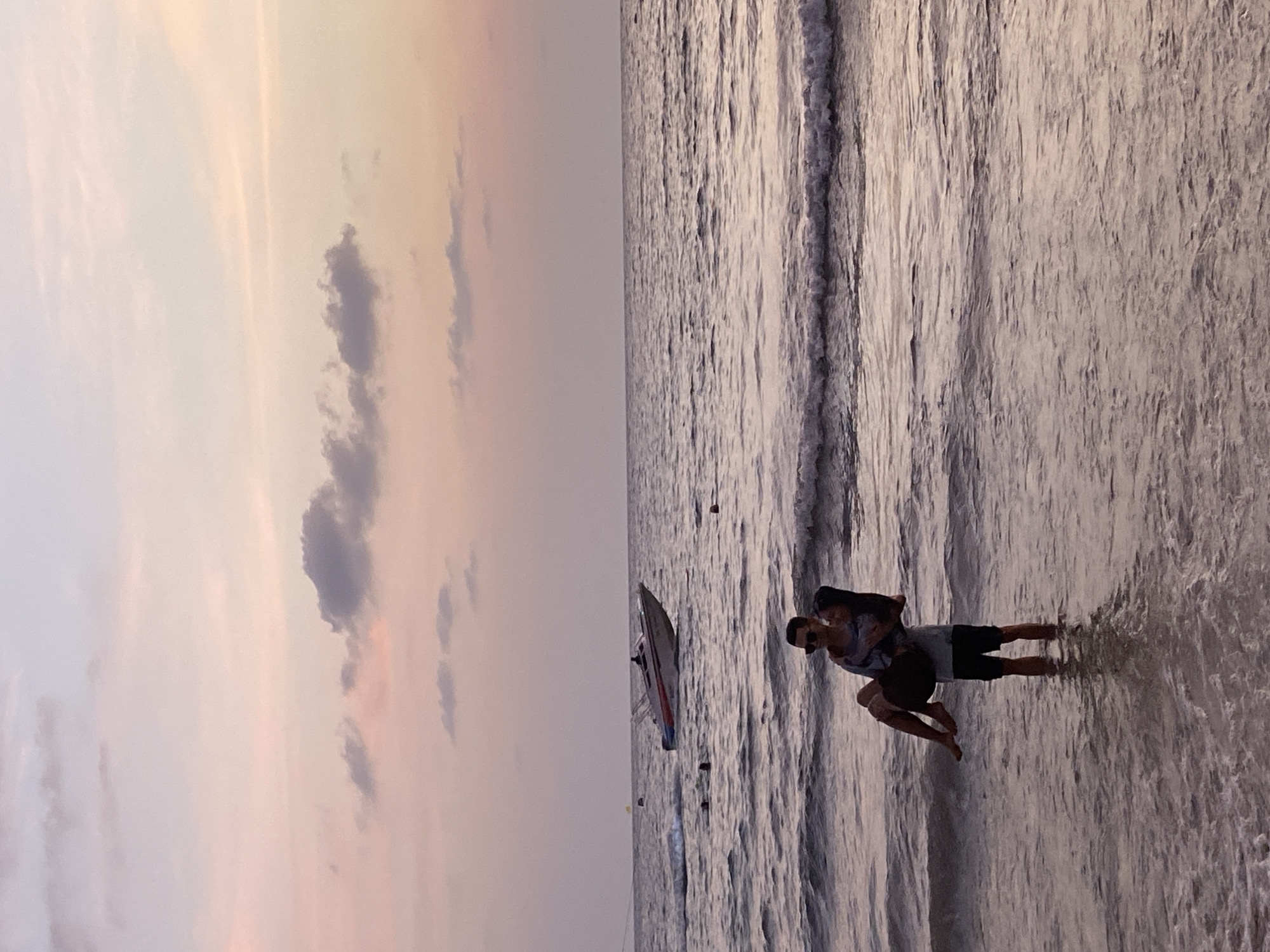 The image captures a scene at Karon Beach in Thailand. The foreground features a man carrying a woman on his back as they stand in the shallow water near the shoreline. They appear to be embracing or enjoying a playful moment. In the background, there's a sailboat floating on the water, and the sky is filled with a soft, muted light, suggesting it is either sunrise or sunset. The beach stretches out, and the water is gently lapping at the shore. The overall atmosphere seems calm and serene.