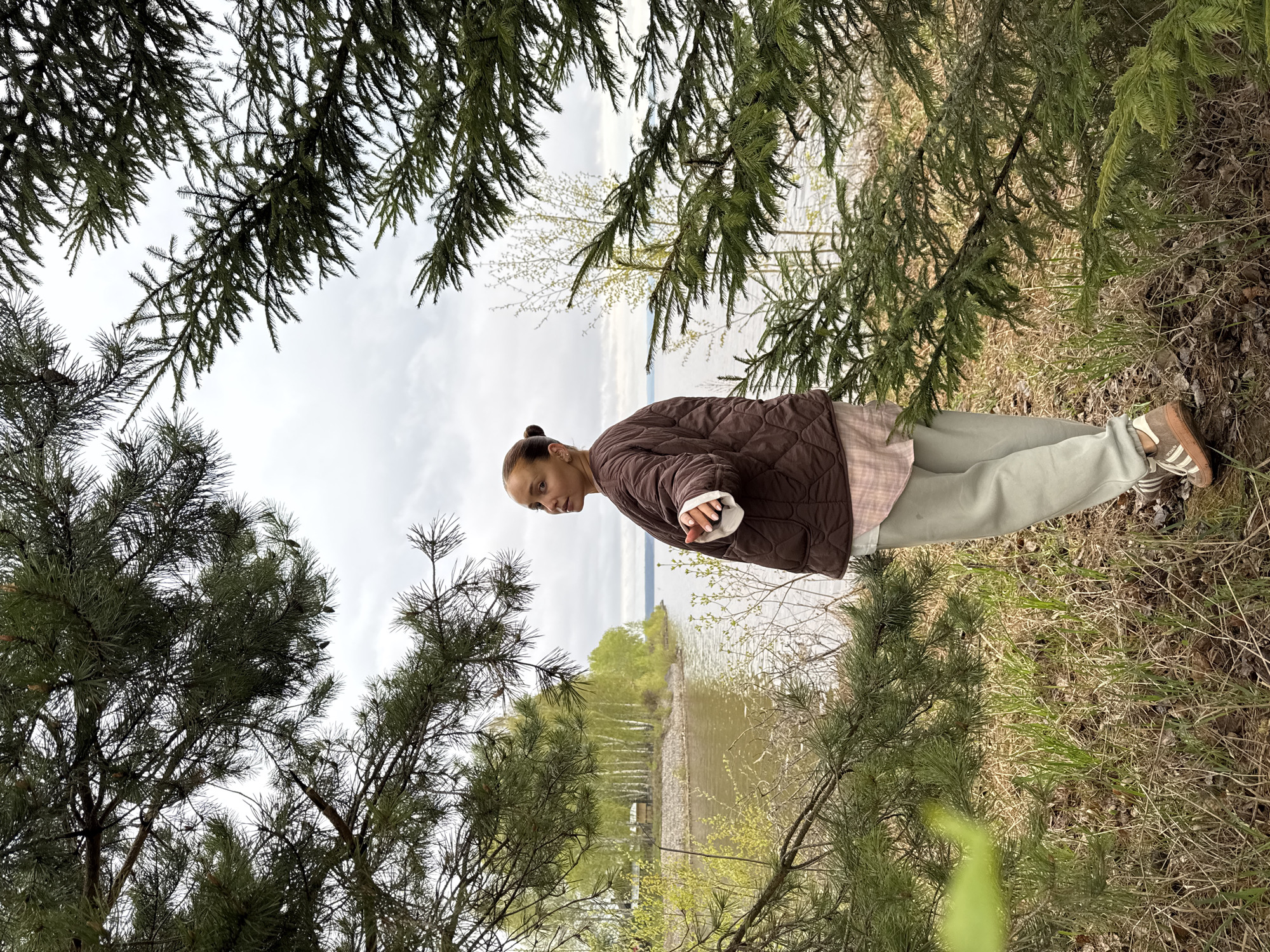 A medium shot, framed by pine tree branches in the foreground, depicts a woman standing near a body of water. The woman, with her hair pulled back in a bun, is wearing a brown quilted jacket over a pink plaid shirt, light green pants, and brown sneakers with cream-colored stripes. She is looking towards the left. The body of water in the background appears to be a lake or river, with a forested shoreline visible across the water. There are various trees and vegetation surrounding the woman. The sky is overcast. The location is Chastyye, Russia.