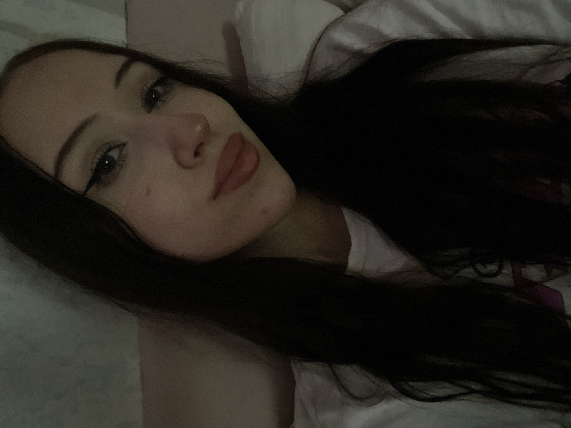 A selfie is taken by a young woman with long, dark hair, wearing a white t-shirt. She has winged eyeliner and neutral-toned lipstick. The background appears to be a dimly lit room with a textured wall and a white pillow. The image does not provide any information to determine the location in Votkinsk, Russia.