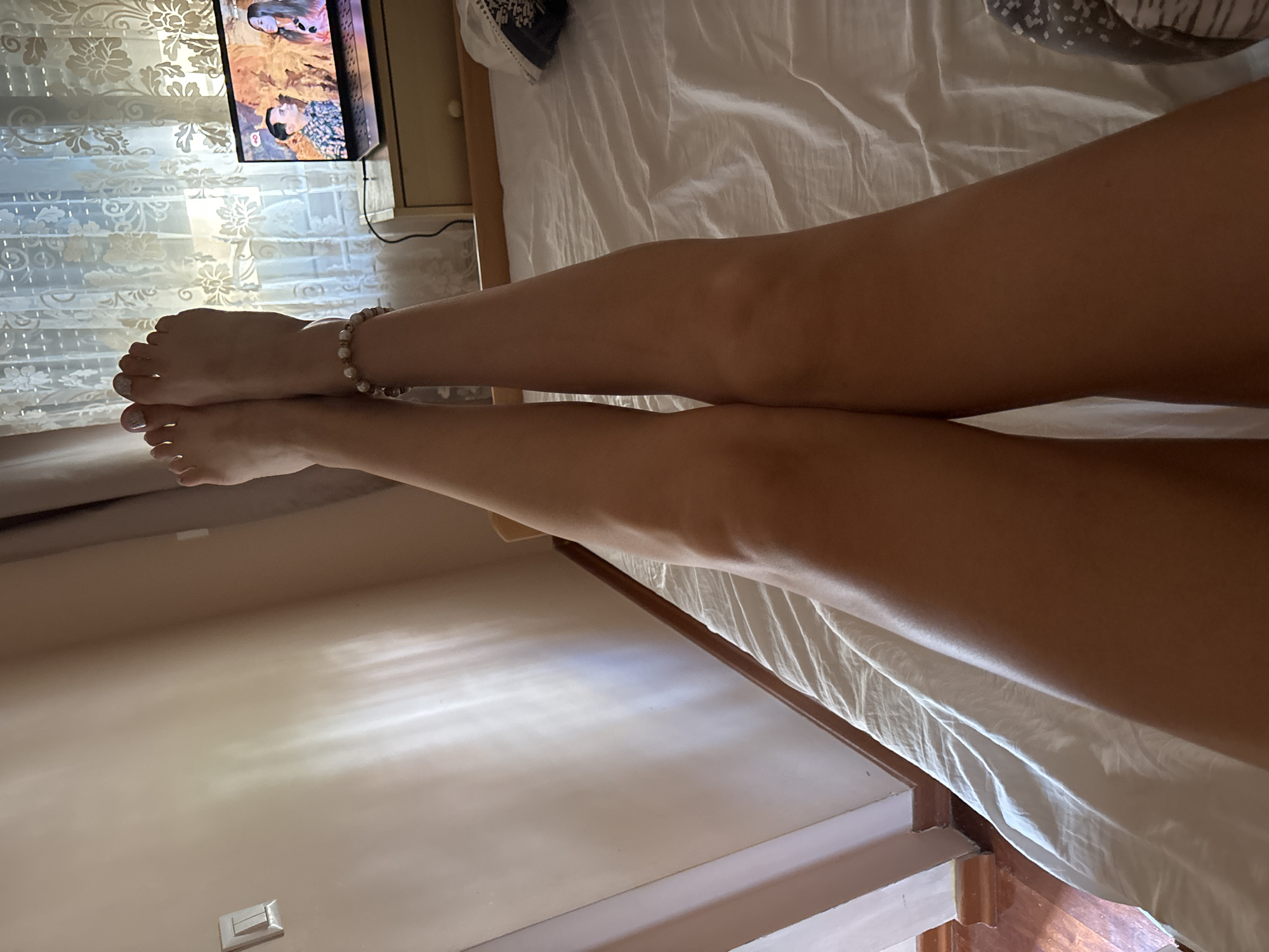 A person's legs, from mid-thigh to toes, are extended upwards and crossed at the ankles, resting on a white-sheeted bed. The person is wearing a beaded bracelet on their right ankle. To the right of the legs, a laptop is open on a bedside table, displaying an image of two people. Behind the laptop, a sheer, patterned curtain is visible. The room appears to be illuminated by natural light casting shadows on the wall. The overall scene suggests a relaxed indoor setting, possibly a bedroom in Budva, Montenegro, given the context of the request.