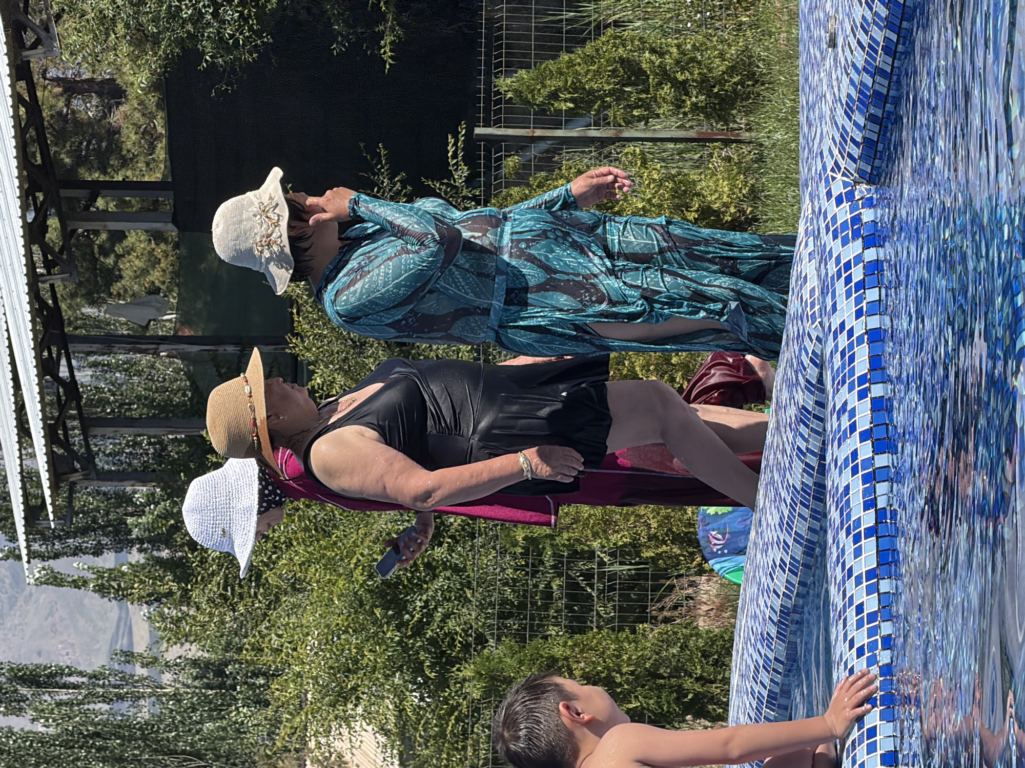 Three women and a young boy are near a tiled swimming pool. The women are wearing hats and swimwear. One woman in a black swimsuit is holding a smartphone and appears to be looking at it. Another woman, wearing a patterned blue and green long-sleeved dress and a light-colored hat, is standing with her hand to her face. The third woman, wearing a pink garment and a white hat, is mostly obscured. The boy, with wet hair, is positioned in the foreground, near the edge of the pool. The background includes lush green foliage, trees, a fence, and a structure with a white roof. The overall setting suggests a sunny day, possibly at a resort or a private residence.