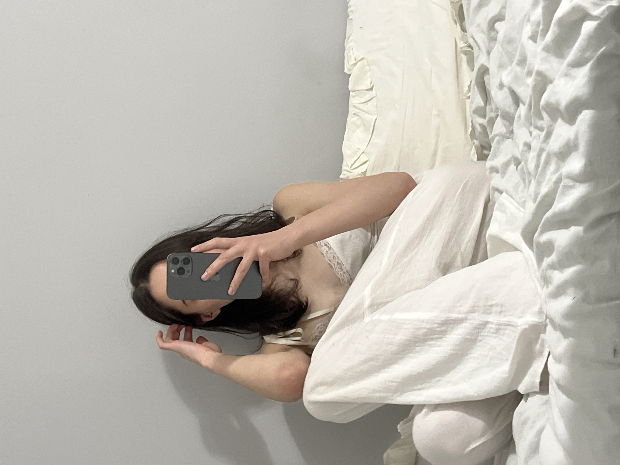 The image shows a young woman taking a mirror selfie in a dimly lit bedroom. She is sitting on a bed, wearing white pajamas and holding a smartphone. Her hair is dark and she has her hand resting on her head. The background is a plain gray wall and the bed is covered with white sheets. The image does not contain any information or clues about the location being Ternopil, Ukraine.