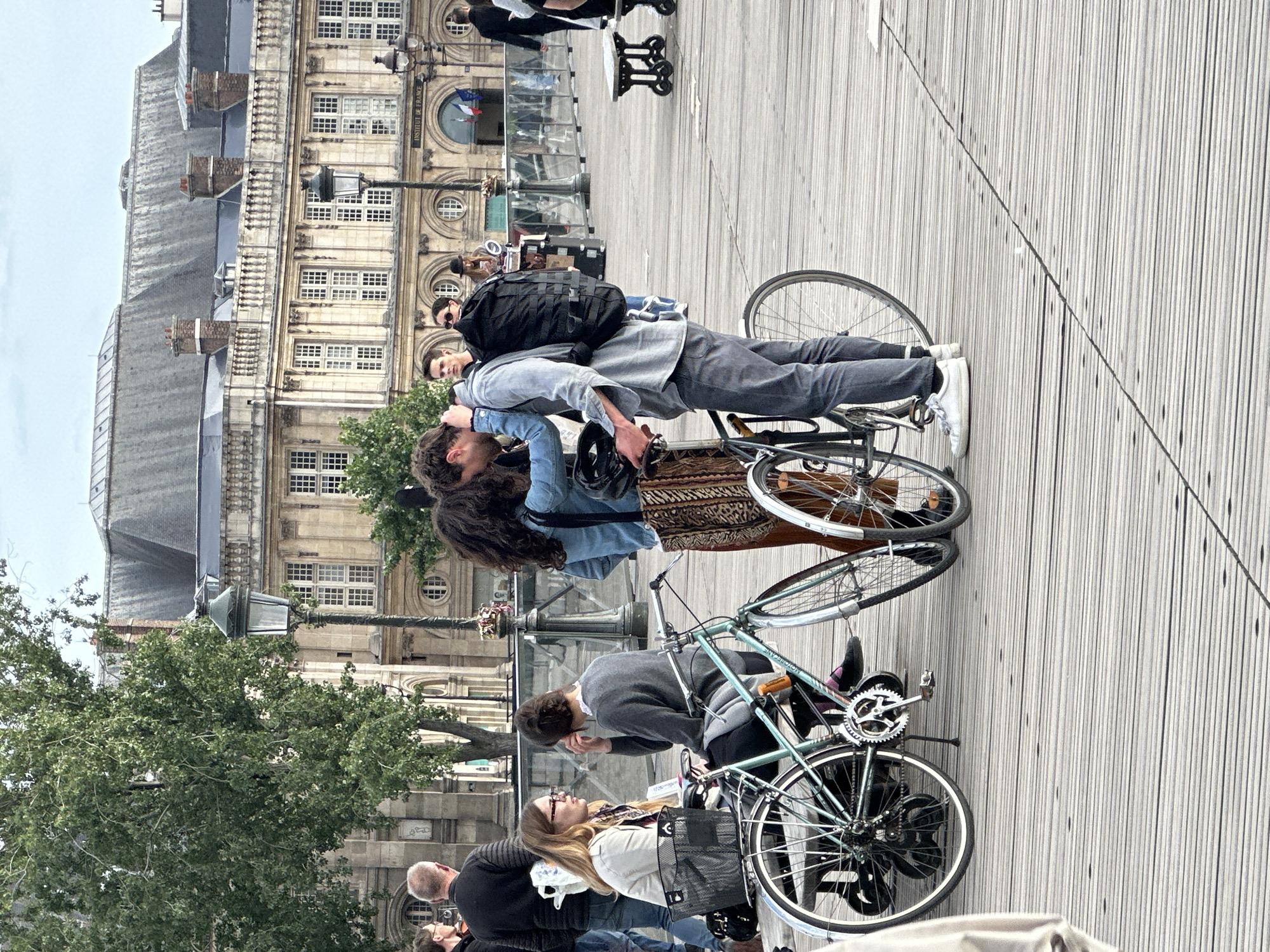 The image depicts a scene in Paris, France, in front of the Institut de France. Two couples are present, seemingly on a date, with each couple positioned near a bicycle. One couple is embracing intimately, while the other couple appears to be preparing to cycle. The setting includes wooden boardwalk-style flooring and a street lamp. People are wearing casual clothing, and the architectural backdrop of the Institut de France is visible in the background.