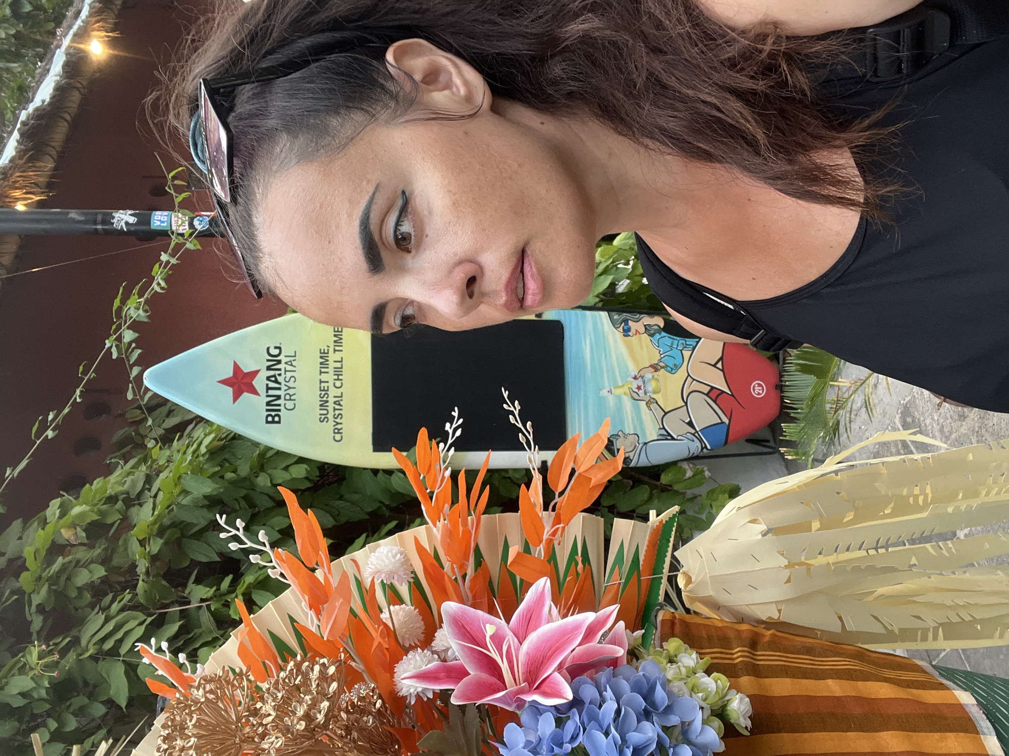A woman with dark hair pulled back in a ponytail and sunglasses resting on her head poses for a selfie in front of a display. She is wearing a black tank top. To her left is a floral arrangement featuring large pink lilies, blue hydrangeas, and orange foliage. Behind her is a surfboard with the Bintang Crystal logo and graphics of people on a beach. The background shows lush green foliage and a rustic building with a thatched roof and string lights. The scene appears to be outdoors, possibly in a market or restaurant area in Denpasar, Indonesia, given the Bintang branding, a popular Indonesian beer.
