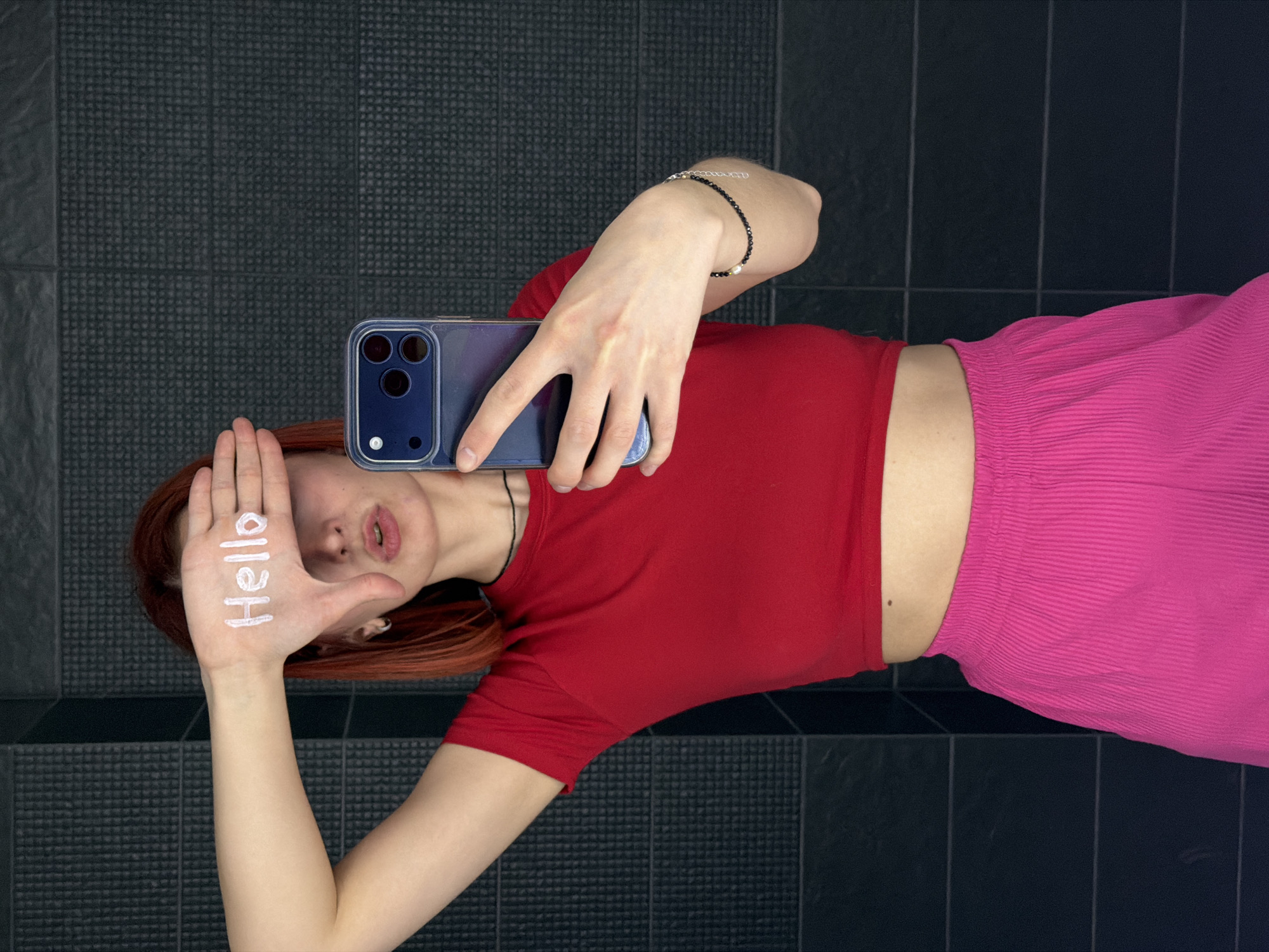 A young woman takes a selfie while lying on a dark tiled floor. She is wearing a red t-shirt and pink pants. She holds her phone in her right hand and covers part of her face with her left hand. The word "hello" is written vertically on her left hand. She has reddish-brown hair and is wearing a black necklace. The location is likely a bathroom or similar room in an unknown location within the city of Chaikovsky, Russia.