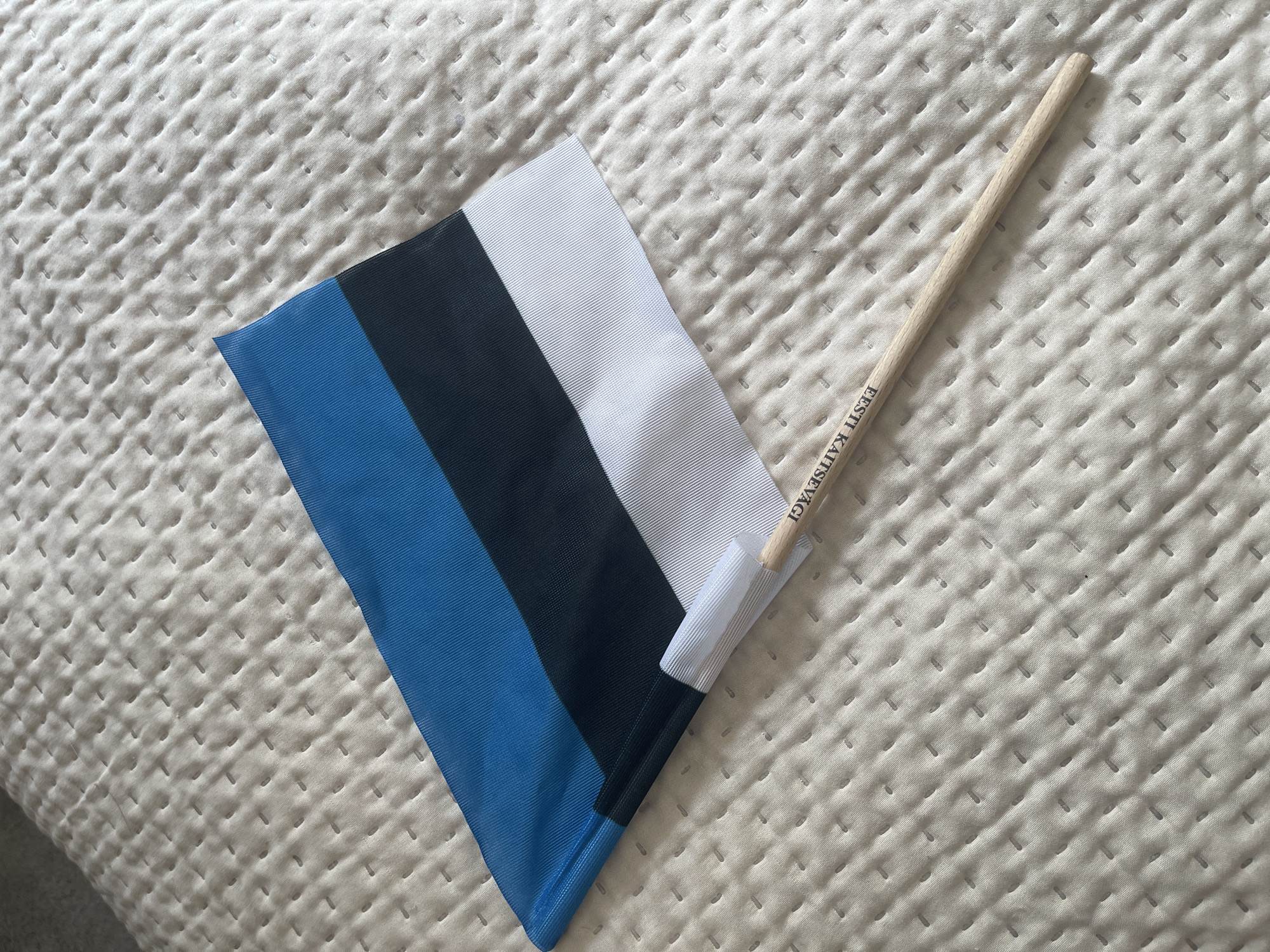 The image shows a close-up, high-angle shot of a small Estonian flag lying on a textured, light-colored surface, possibly a quilted blanket or mattress. The flag is positioned diagonally across the frame, with the blue stripe at the top, followed by black, and then white at the bottom. A thin, wooden dowel serves as the flag's pole, and it extends out of the bottom left corner of the flag. Black text, partially visible and oriented along the dowel, reads "EESTI KAITSEVÄGI," which translates to "Estonian Defence Forces." The background surface exhibits a regular pattern of stitched indentations. The scene is lit from above, casting soft shadows. No people or specific city locations are depicted.