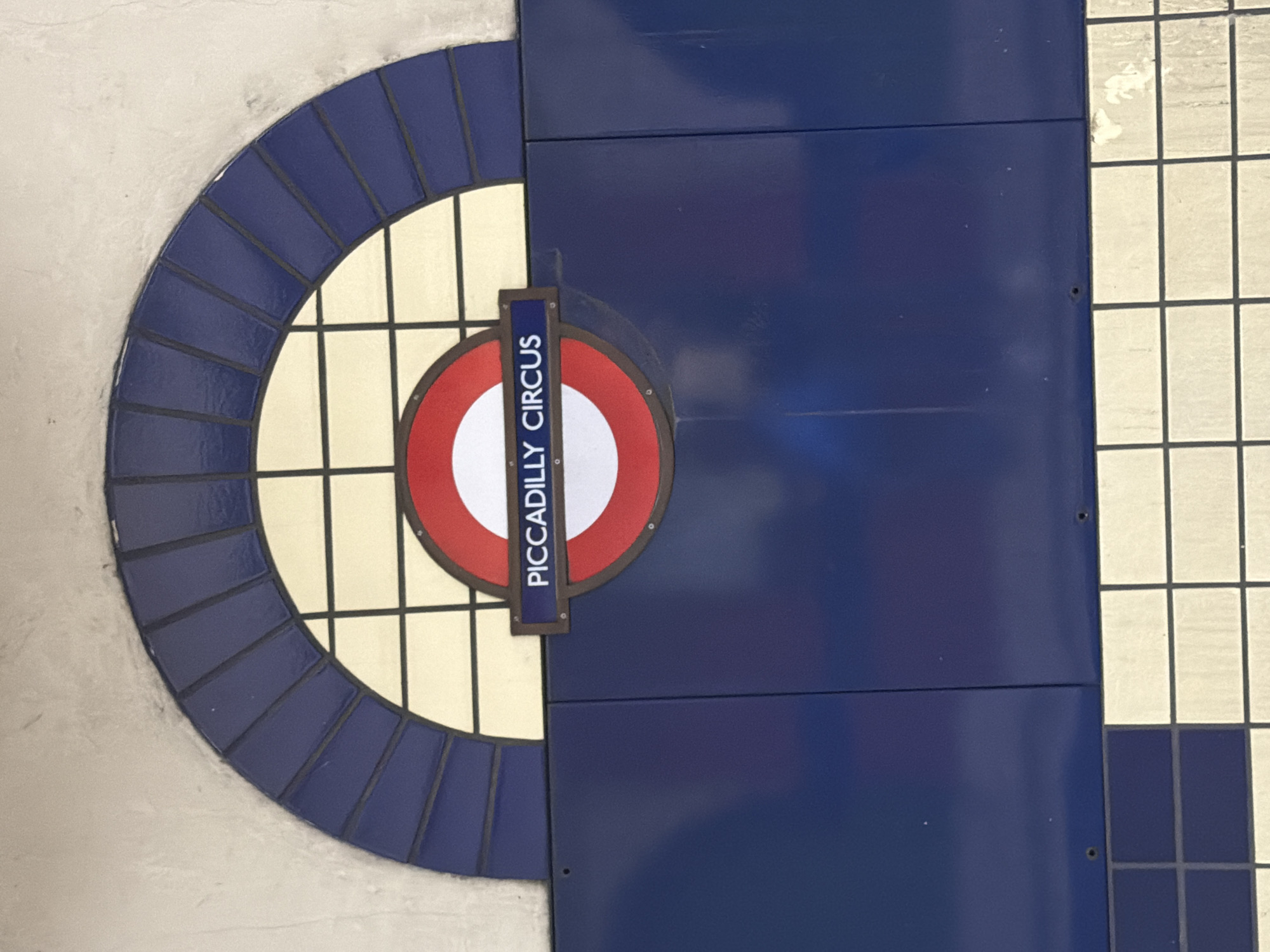 The image shows a section of the tiled wall at the Piccadilly Circus London Underground station. The wall features a semicircular design with a dark blue tiled frame and a grid pattern of cream-colored tiles within. A circular London Underground sign bearing the name "Piccadilly Circus" is mounted over the design, partially obscuring the grid. To the right of the sign and design are dark blue rectangular tiles extending to the edge of the frame. A section of white and blue square tiles can be seen to the right of the blue rectangular tiles.