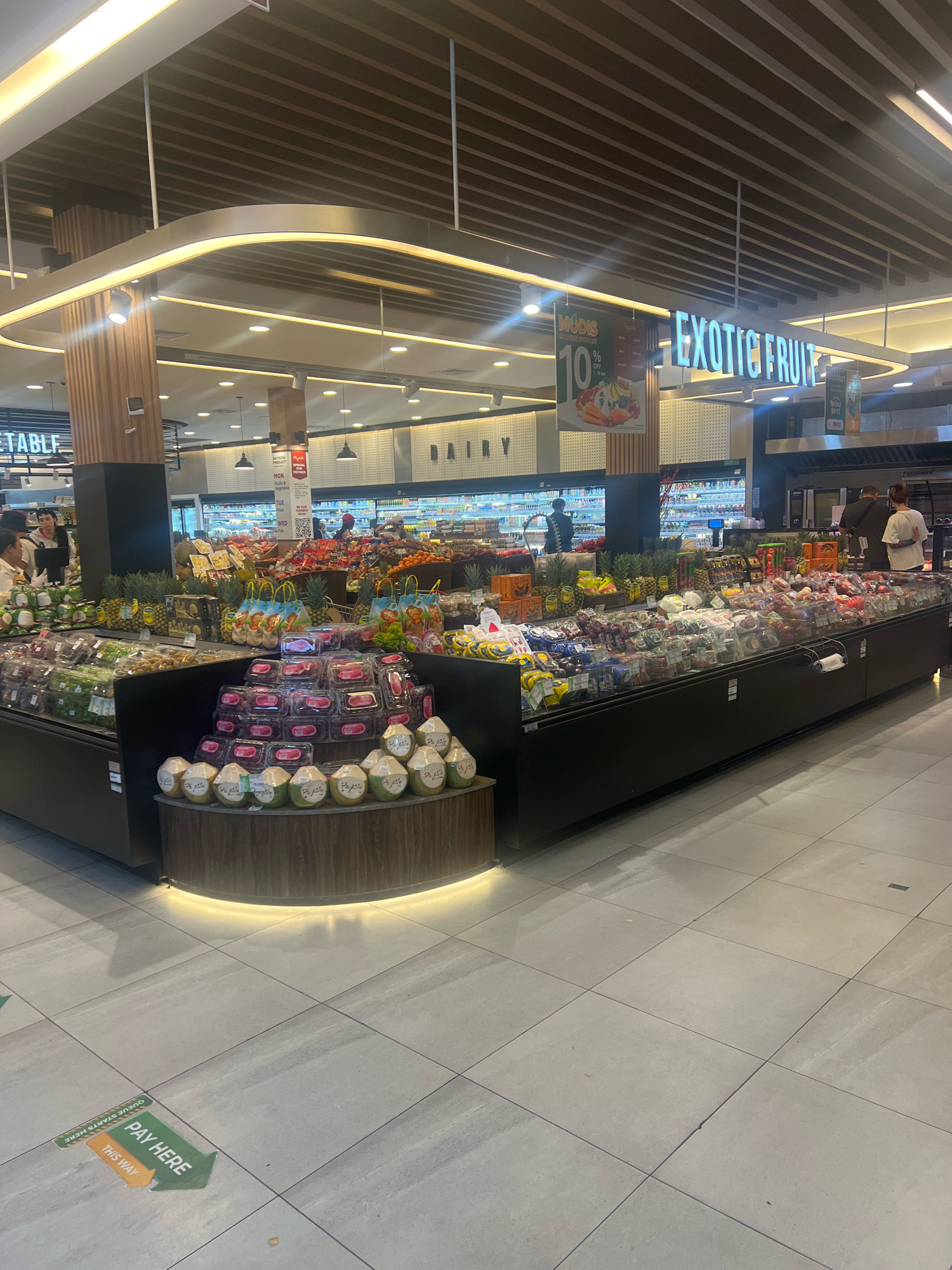 An interior view of a grocery store in North Kuta, Indonesia, features a well-lit produce section with a variety of fruits and vegetables. In the foreground, a tiered display showcases an abundance of packaged fruits like lychees and strawberries, alongside whole coconuts arranged on a curved wooden base. A large refrigeration unit filled with produce is visible in the background, with several shoppers browsing the aisles. Overhead, a modern ceiling with wooden slats and recessed lighting illuminates the space. A prominent sign displaying "EXOTIC FRUIT" is suspended from the ceiling, and a promotional banner advertising "MODIS 10%" is also visible. On the floor, green and orange directional arrows point towards a "PAY HERE" station, indicating a designated checkout area.