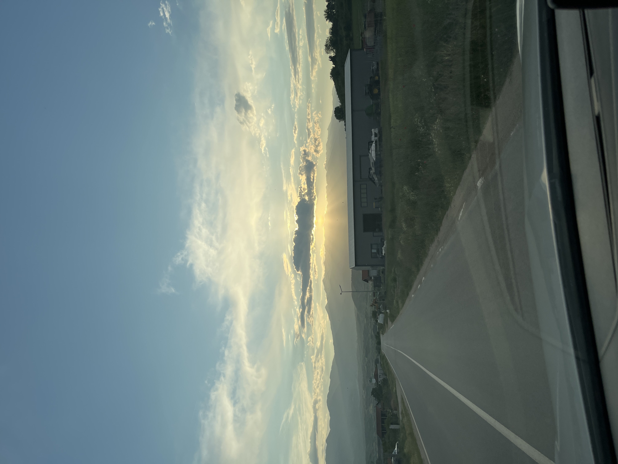 The image shows a view from a moving vehicle, likely a bus or car, heading towards what appears to be a setting sun obscured by clouds. In the distance, a line of mountains is visible. A two-lane road with a white dividing line stretches ahead, with buildings and vegetation along the roadside. The location is suggested to be near Baldevo, Bulgaria. The vehicle's window is partially visible on the right side of the image, adding a sense of depth and indicating the perspective is from inside the vehicle. The scene evokes a feeling of travel and possibly sunset.