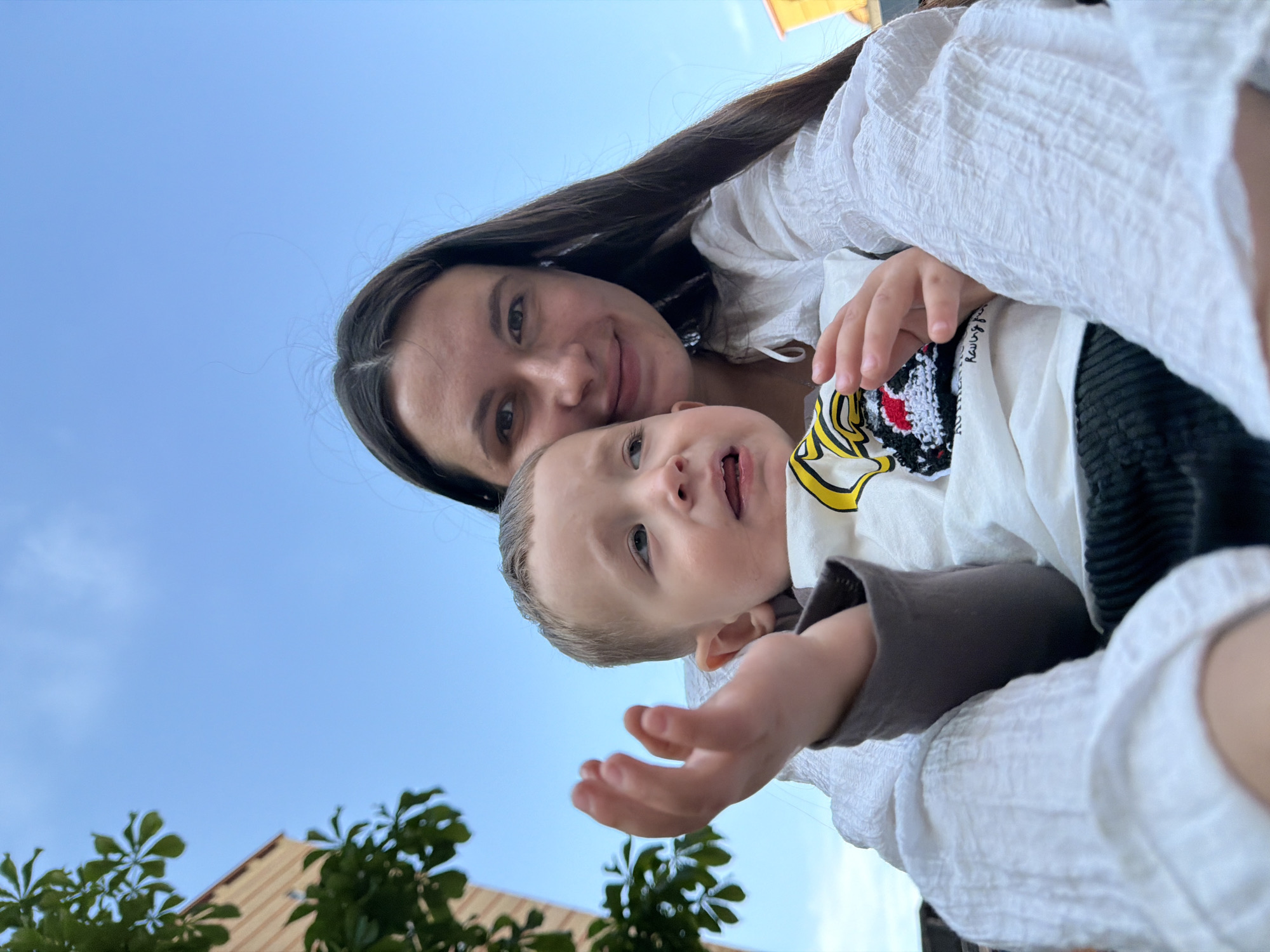 A close-up, low-angle selfie shows a woman and a baby. The woman, with long, dark hair, smiles warmly at the camera. She is wearing a white, textured garment. Next to her, partially obscured, is a baby looking towards the camera with an open mouth, appearing to be mid-cry or mid-laugh. The baby is wearing a white t-shirt with a colorful cartoon character and yellow accents. They are both outdoors, with a clear blue sky as the background. In the lower part of the frame, blurred green foliage and a portion of a building with a beige roof are visible, suggesting a park or garden setting. The lighting suggests it is daytime, likely late afternoon or early evening given the soft light. The overall mood is intimate and affectionate, capturing a candid moment between the two. The text "Rawrg?" is visible on the baby's t-shirt. The location context is Vniissok, Russia.
