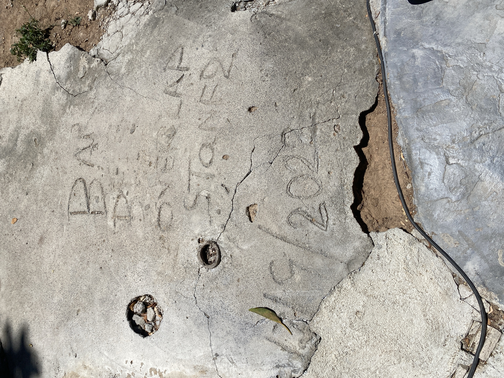 The image shows a cracked concrete surface with writing etched into it. The writing appears to be in English and includes the words "BAR" and "STONE". Below these words, there is what looks like a date, possibly "2023". There is also a circular indentation with debris inside, a loose leaf, and some exposed soil with a black cable running across it. The scene is outdoors, under sunlight, with shadows cast on the concrete. There is no indication of the city of Baan Lamai, Thailand in the image itself.