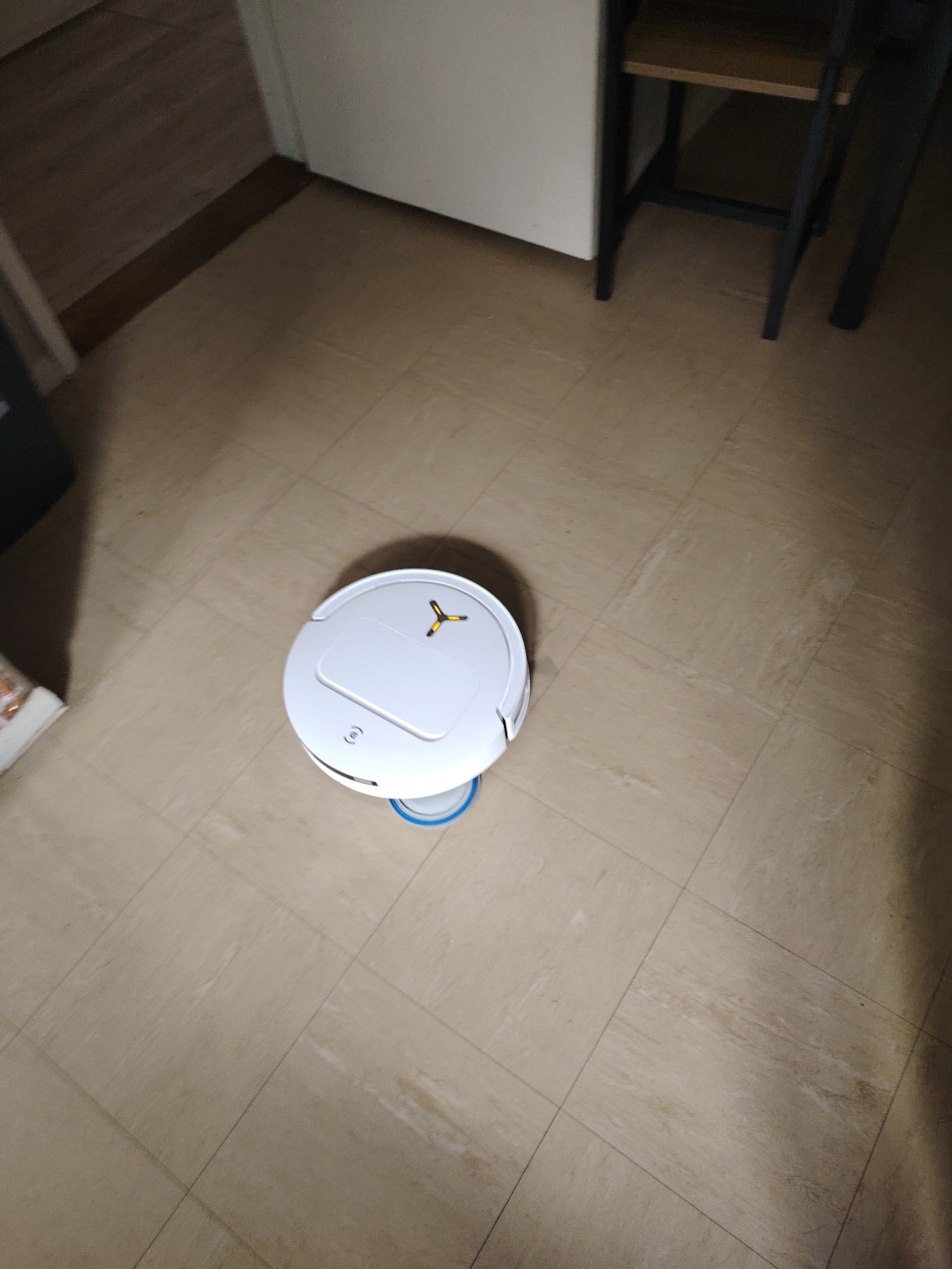 A white, circular robotic floor cleaner, featuring a blue-ringed base and a yellow three-pronged symbol on its upper surface, rests on a light beige tiled floor. The tiles display subtle streaked patterns with visible grout lines. To the upper-left, a wooden door or cabinet and a light-colored wall with a base shadow are present. The upper-right reveals dark metal furniture legs and a wooden shelf, indicating a piece of furniture like a chair or table. The area is dimly lit, with shadows concentrated on the right. No persons are depicted. This scene is situated in Huntingdonshire, United Kingdom.