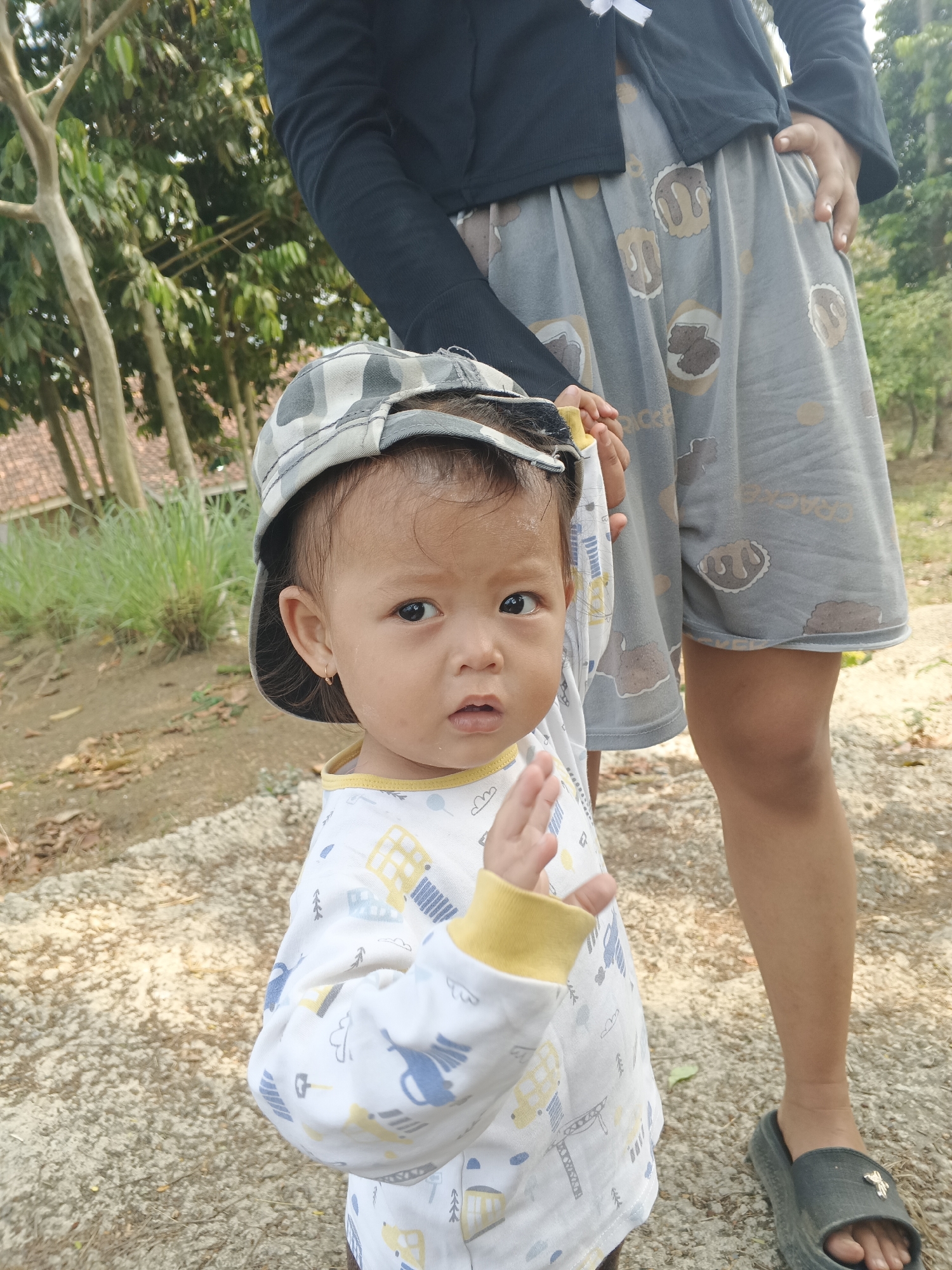 The image shows a close-up of a young child wearing a camouflage baseball cap and a long-sleeved shirt with a yellow collar. The child's left hand is raised, palm facing forward. Behind the child, an adult's legs are visible, clad in grey patterned shorts and dark sandals. The background features trees and vegetation, suggesting an outdoor setting, likely in a residential area or park. The ground is a mix of dirt and gravel. The lighting indicates it is daytime, possibly late afternoon due to the warm tone of the sunlight. The scene appears to be a casual moment captured outdoors, potentially in Sukabumi, Indonesia, given the context provided.