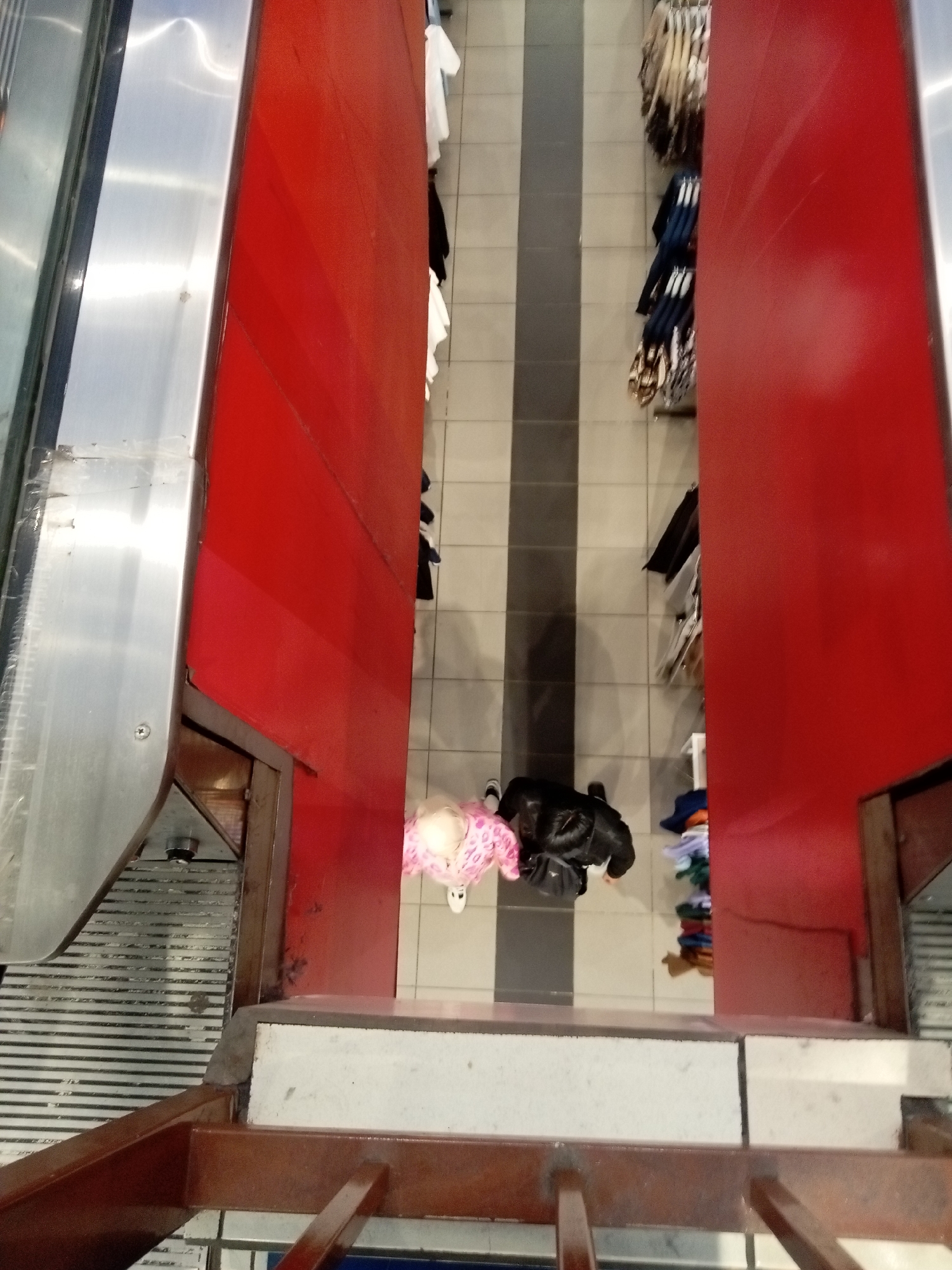 This is a high-angle, full shot of a department store interior in Batam, Indonesia. The scene captures the entrance to a clothing section, with two people visible. On the left, a person wearing a pink and white patterned outfit stands near a red display. To the right, a person in a black jacket and black pants is visible. The store features tiled flooring with grey and white stripes, and clothing racks displaying various garments, including what appear to be shirts and pants. The environment suggests a commercial setting within a shopping mall or department store.
