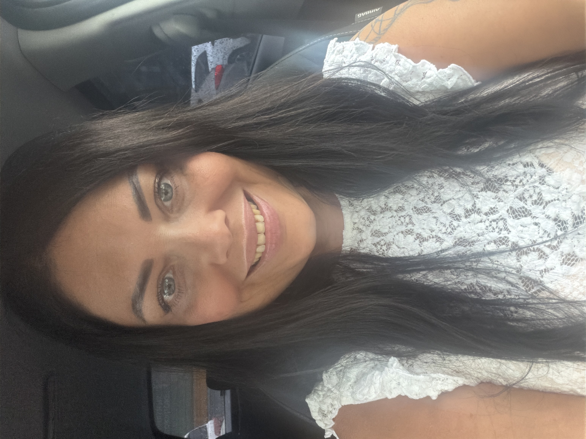 A woman with long, dark hair and light eyes is smiling at the camera. She is wearing a white lace top. The setting appears to be inside a car. There is no information to determine if the image was taken in Murmansk, Russia.