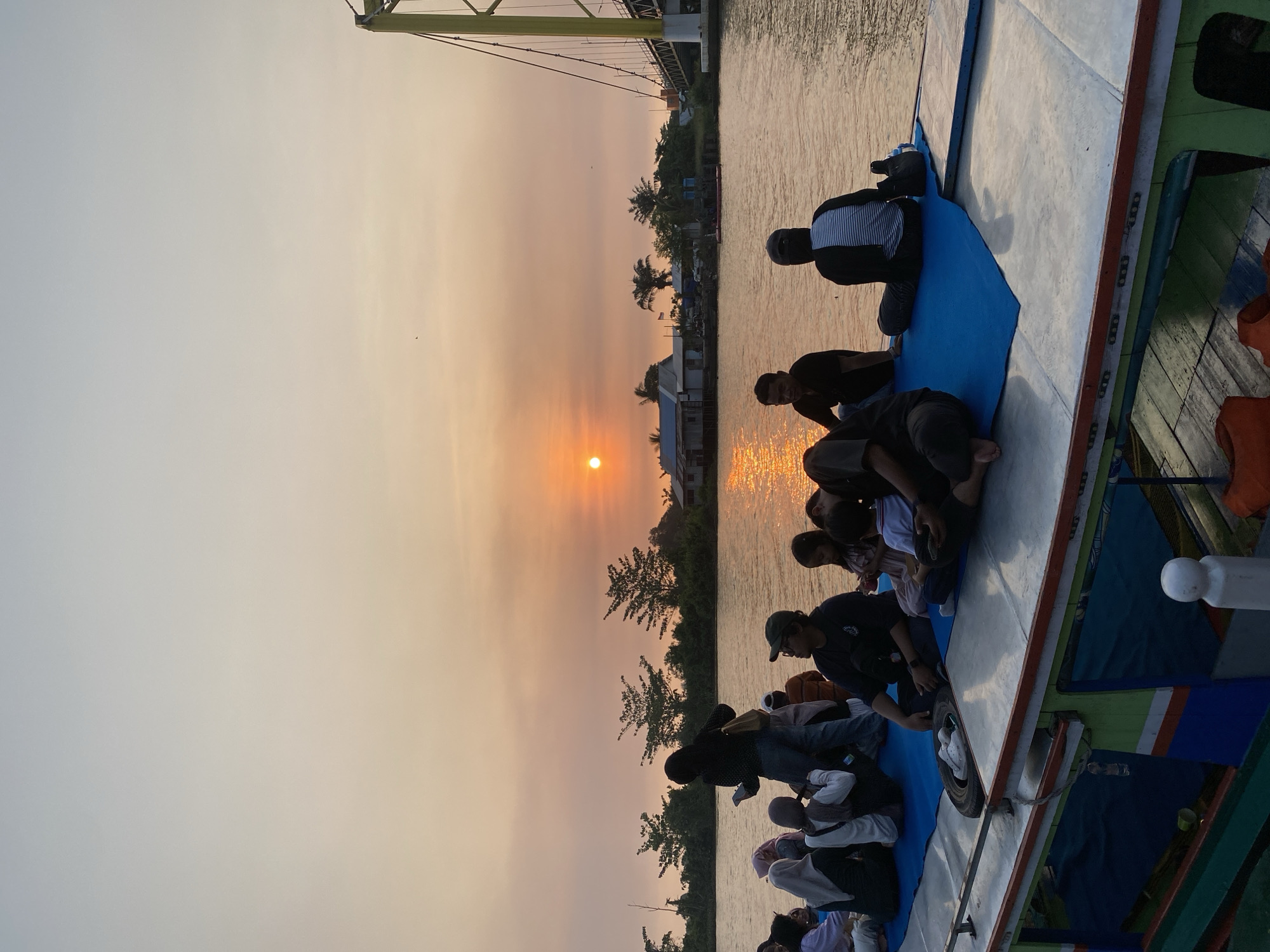 The image depicts a sunset scene over a body of water, likely a river, in Sungai Pitung, Indonesia. A group of people are seated and standing on the deck of a boat. The boat is painted in vibrant colors with green, blue, and orange accents. The people are diverse in age and gender, with some children present. They are casually dressed and appear to be enjoying the view of the sunset.

The sky is a soft gradient of orange and pale blue, with the sun positioned low on the horizon, casting a warm glow on the water. In the background, there are trees and a few buildings on the shore, suggesting a rural or semi-urban setting. A yellow suspension bridge is visible on the right side of the image, spanning the water. The overall atmosphere is serene and peaceful, indicative of a relaxed outing.