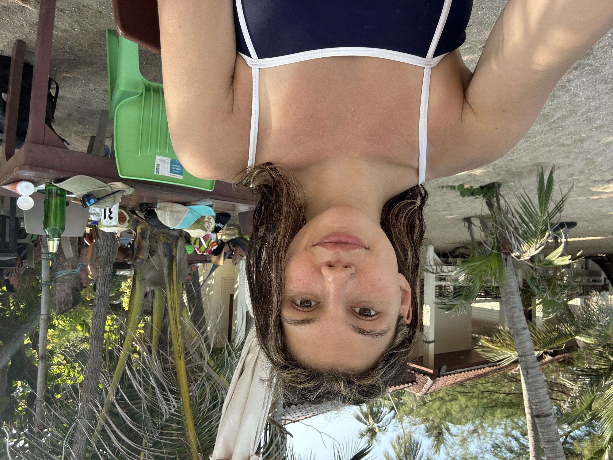 The image is a selfie taken by a young woman with light brown, shoulder-length hair and brown eyes. She has a fair complexion and a piercing in her left nostril. She is wearing a dark blue swimsuit with white straps. The background consists of a tropical setting with palm trees and a building. There is also a table with various items on it, including a green bottle and a hat. The image appears to be taken outdoors during daylight.
Based on the prompt mentioning "Baan Chaweng Noi, Thailand," it is likely the image was taken in that location, though this cannot be definitively confirmed solely from the visual information.