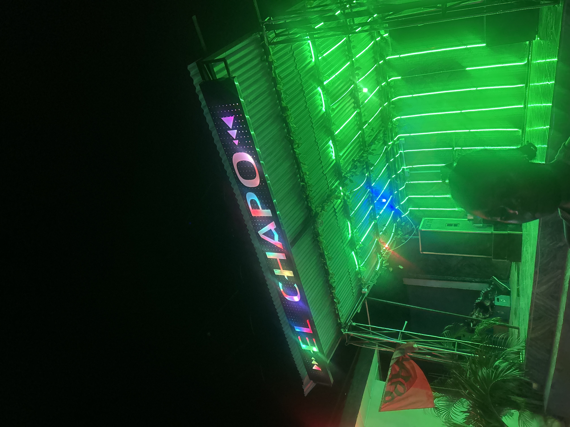 This image shows a nighttime exterior view of a venue with a prominent LED sign reading "EL CHAPO" in rainbow colors. The sign is mounted on a corrugated metal roofed structure, which is heavily decorated with vertical green LED lights. To the left, a red flag with a black symbol is visible, and to the right, there are black speaker boxes. The foreground is dimly lit, with a person's silhouette visible on the right side of the frame. The background is dark, suggesting it is nighttime in the city of Masaka, Nigeria.