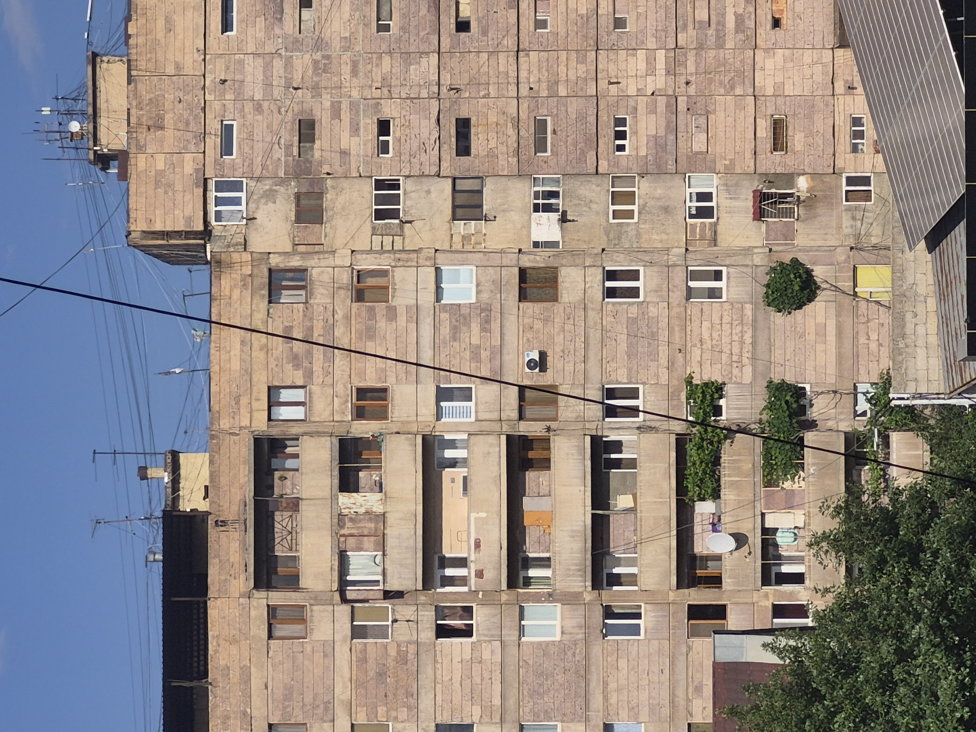This media file depicts a tall, multi-story residential building, characteristic of post-Soviet architecture, situated in Yerevan, Armenia. The building's facade is constructed from numerous light-colored concrete panels, presenting a somewhat worn and utilitarian aesthetic.
The building is densely covered with a variety of windows, reflecting individual modifications over time. These range from modern white-framed windows to older wooden or darker-framed ones, some open, some closed, and some revealing curtains or blinds. Several balconies are present, some enclosed with various materials and others open, occasionally showing stored items or glimpses of daily life.
The exterior is crisscrossed by a network of black electrical wires and cables, running both horizontally and vertically. Visible utilities include at least two white satellite dishes and two air conditioning units. The rooftop features multiple antennas and communication equipment set against a clear blue sky.
Natural elements include a small, bright green bush growing from one of the building's upper sections, and a larger green tree partially obscuring the lower right portion of the building. A prominent, angled structure, possibly solar panels or a unique roof design, is visible in the upper right quadrant of the image.
The scene is captured during clear, sunny daylight hours, indicated by the bright, even lighting and cloudless sky. No people or specific activities are visible, and there is no discernible text anywhere in the image. The overall impression is that of a well-used and adapted urban dwelling.