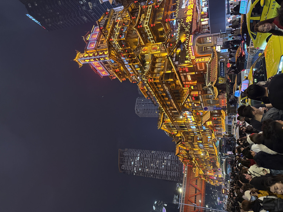 The image displays a nighttime urban scene in Yuzhong District, Chongqing, China, featuring the illuminated Hongyadong architectural complex. This multi-tiered structure, built in traditional Chinese style, occupies the right side of the frame and is extensively lit with golden lights and numerous red lanterns. Neon signs displaying Chinese characters, including "洪崖洞," are visible on its upper facades. In the background, several modern high-rise buildings pierce the dark sky, one appearing partially constructed or undergoing renovation. A red-orange bridge structure is visible in the mid-ground on the left. The street level is densely populated with a large crowd of pedestrians, predominantly adults, dressed in casual attire, standing and congregating. The street itself is busy with vehicular traffic, including multiple yellow taxis, other cars, and at least one bus. The entire scene is brightly illuminated by artificial lights emanating from buildings, streetlights, and vehicles. No specific individual interactions are discernible within the crowd; individuals appear to be generally observing the environment.