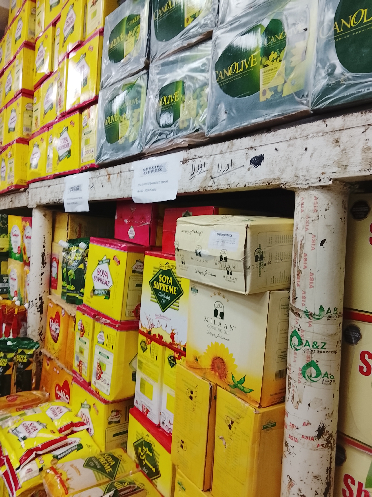 The image shows a shelf in what appears to be a retail store in Gulgasht Colony, Pakistan. The shelves are heavily stocked with various cooking oils, including "CANOLIVE," "SOYA SUPREME," and "MILAAN" brands. The oils are packaged in boxes and plastic containers. There are signs indicating "SPECIAL OFFER" for some products. The shelves and support columns are aged, with visible wear and tear. There is writing in Urdu on the shelf. The support column is covered with "ASIA" advertisements.