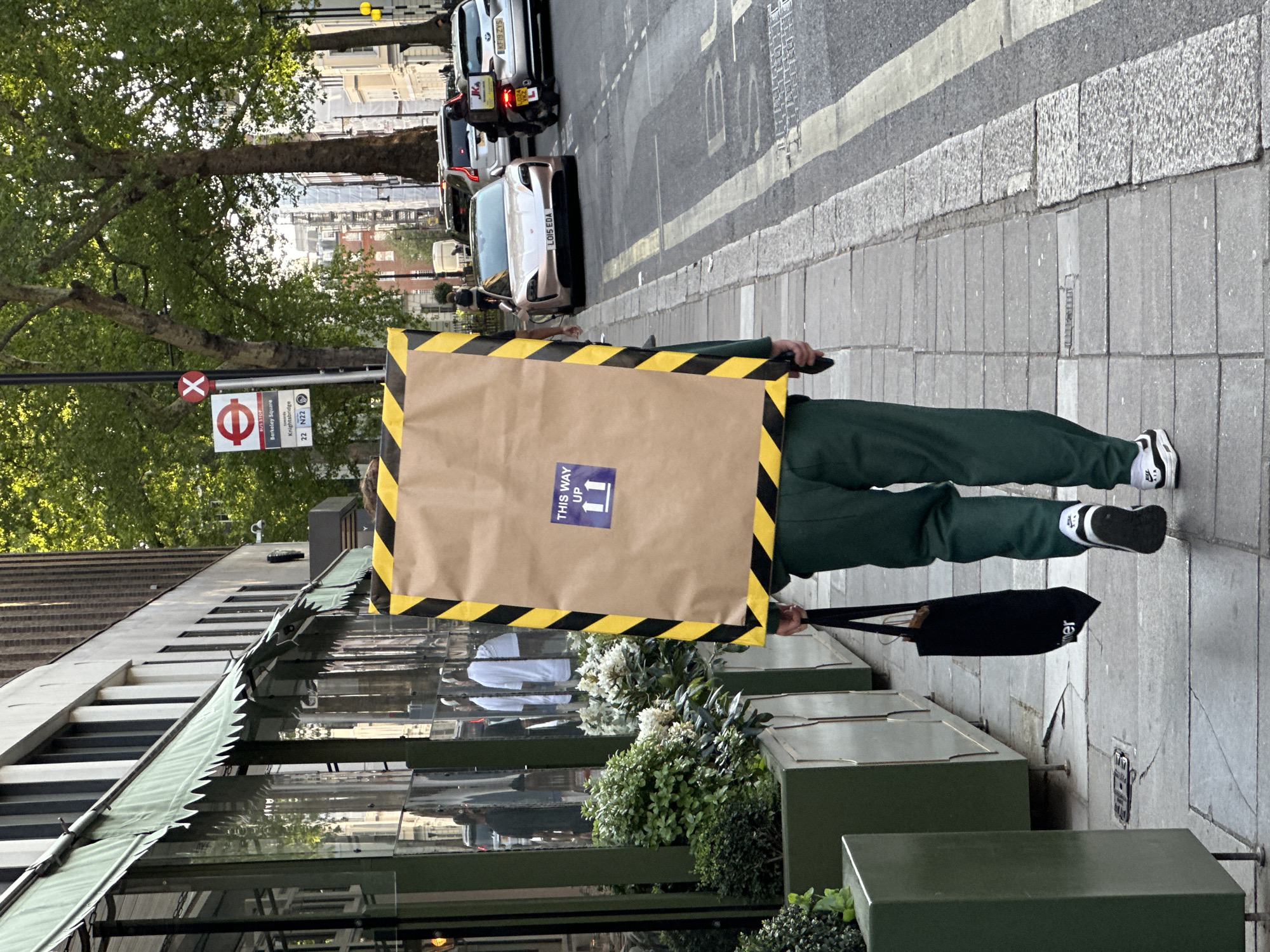 A person in a green tracksuit, white socks, and black/white sneakers walks along a sidewalk. The person holds a sign framed with yellow and black caution tape that reads "THIS WAY UP." In their other hand, they carry a black tote bag. The scene appears to be a street in London, United Kingdom, as evidenced by the bus stop sign indicating "Berkeley Square, Knightsbridge" and bus routes "22 N22." Several cars are visible in the street, including a silver car with the license plate "L015 EDA" and another vehicle with the obscured plate "KFIS ZYS." The sidewalk is made of gray stone, and a building with an awning is on the left side of the frame. There are trees and other foliage adding to the urban streetscape.
