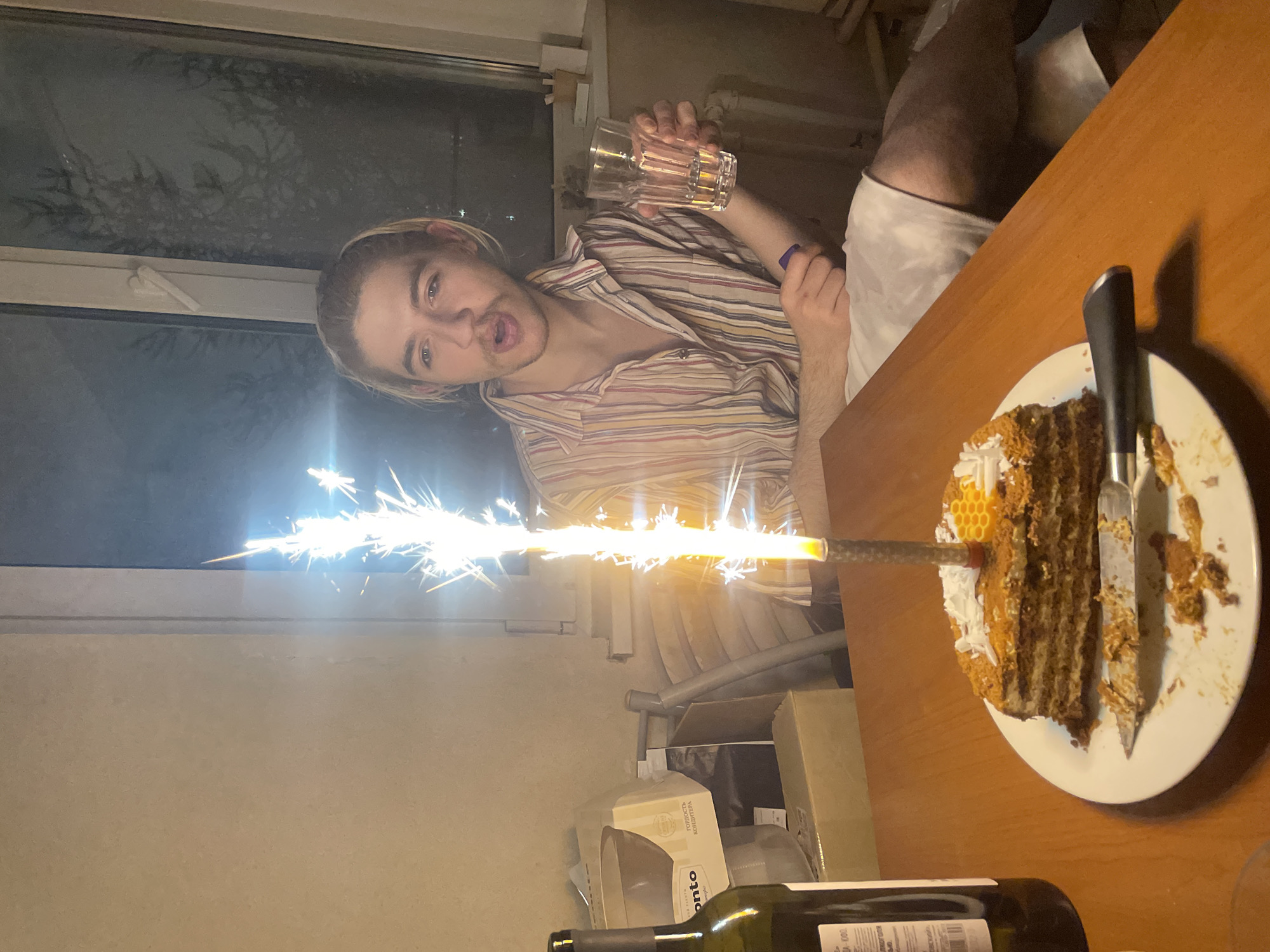 The image depicts an indoor celebratory scene at night, likely in Moscow, Russia. A young man, with blonde hair tied back and light facial hair, is featured prominently. He wears a striped shirt and holds a clear glass, looking directly at the camera with a playful expression, his tongue slightly extended.
In the foreground, on a wooden table, a partially eaten multi-layered cake sits on a white plate. A bright sparkler candle, emitting a cascade of sparks and light, is stuck into the cake, illuminating the area. A knife is also present on the plate, with cake residue. The cake itself appears to be a honey cake or similar, garnished with white shavings and what looks like a small decorative honeycomb. A green glass wine bottle is visible in the bottom foreground.
The background features a dark window, revealing silhouettes of trees against the night sky. The room is casually furnished, with various boxes and other items visible, suggesting a home environment. On one visible cardboard box, the word "ONTO" is printed, along with Cyrillic text that appears to read "Гордость Декора" (Pride of Decor). The overall mood is informal and celebratory, underscored by the man's lighthearted expression.