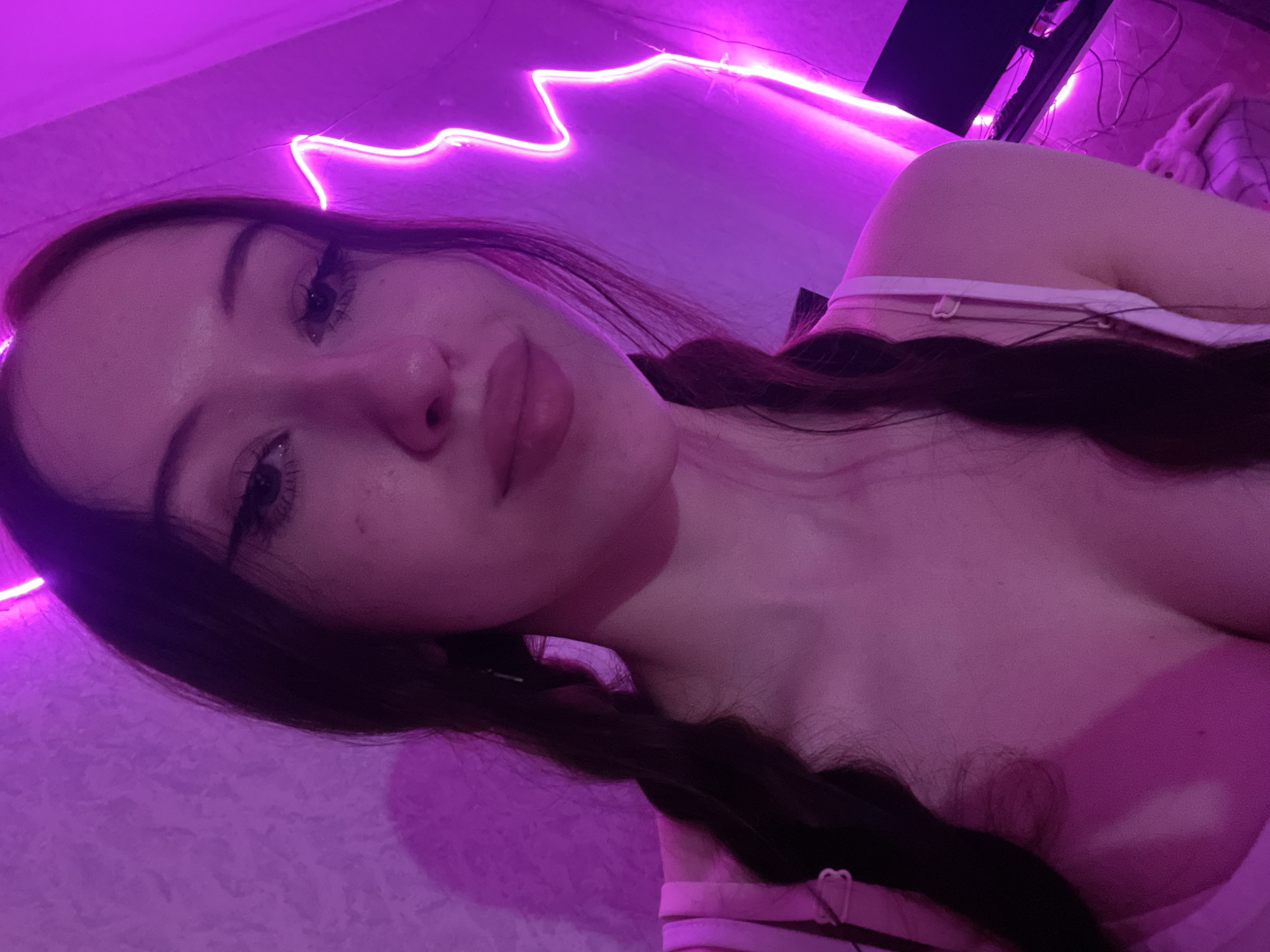 The image features a young woman in a close-up self-portrait, positioned in an indoor setting located in Votkinsk, Russia. She has long, dark hair styled into two braids. Her fair complexion shows visible freckles. She wears a light-colored tank top with visible straps, revealing cleavage. A small, dark pendant on a thin necklace rests on her chest, and a heart-shaped detail is visible on one shoulder strap. The scene is bathed in pervasive magenta and purple light, originating primarily from a visible lightning bolt-shaped neon sign on the right background. A textured wall is behind her, and to the right, a portion of a desk with a black monitor and cables is visible beneath the neon light. A plush object and patterned fabric are also discernible in the lower right background. The woman is looking directly towards the camera.