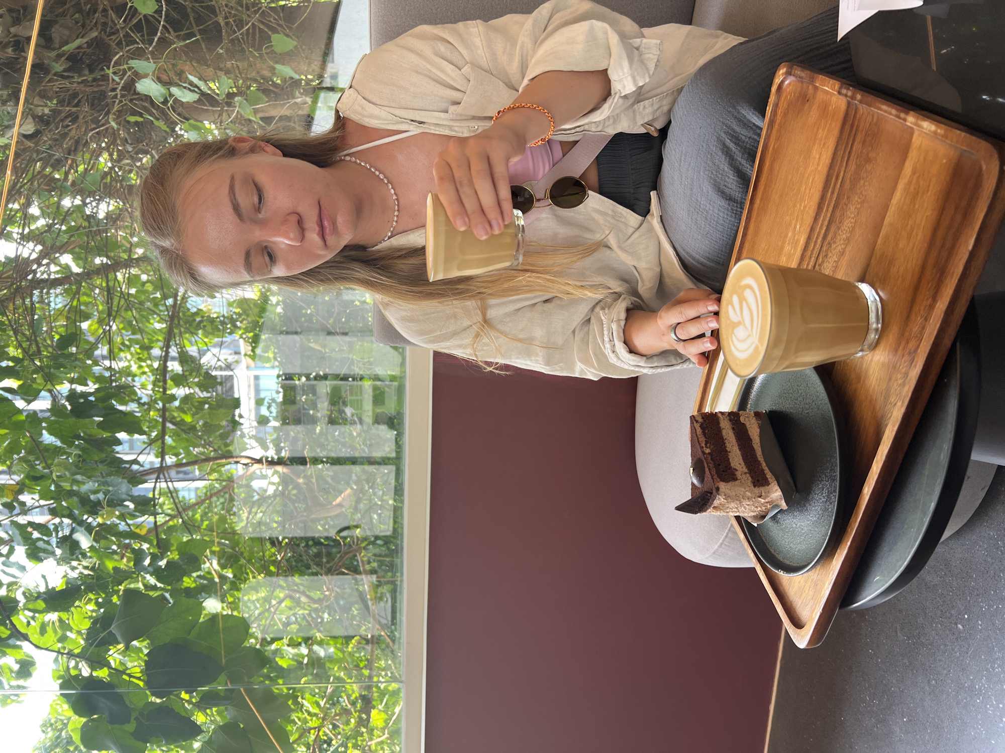 A woman in Bangkok, Thailand, pours coffee at a cafe. She sits in a plush gray chair, dressed in gray pants, a pink top, and a beige overshirt. A wooden tray holds a cup of latte with latte art, a slice of chocolate cake on a dark plate, and a second cup of coffee she is pouring. Her sunglasses rest between the two cups. A string of pearls adorns her neck, and a bracelet decorates her wrist. Outside the window, lush green trees dominate the view.