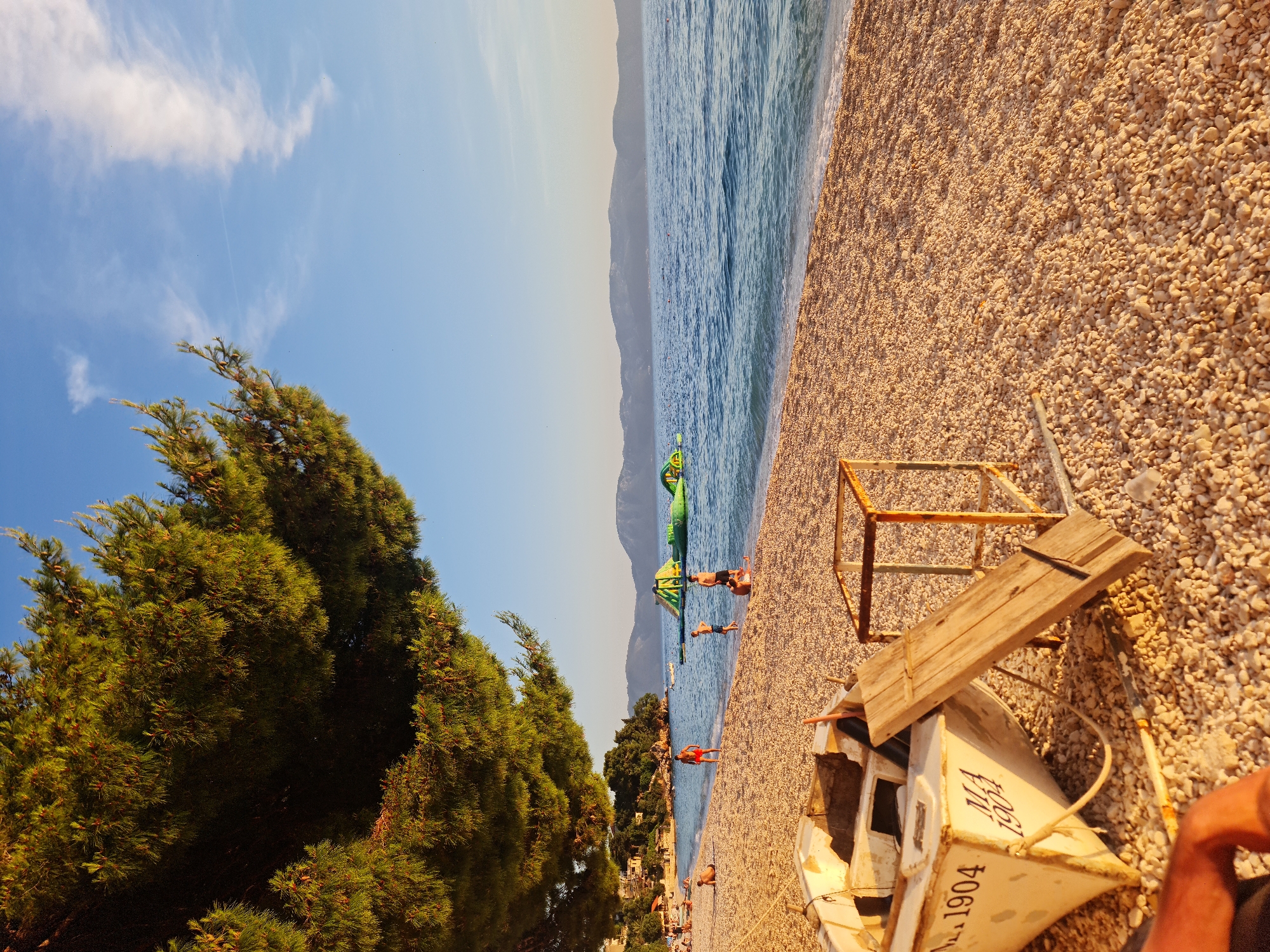 A serene beach scene unfolds in Općina Gradac, Croatia, under a clear blue sky with wispy clouds. The foreground is dominated by a white, weathered boat, its hull marked with "MA 1904". Beside it rests a wooden plank and a rusty metal frame, both appearing discarded. The beach itself is a carpet of small, light-colored pebbles, gently meeting the calm, blue sea.
In the middle ground, several individuals are scattered across the sand and wading in the shallow water. Two young boys, one in blue shorts and the other in dark shorts, are visible near the water's edge. A man in red shorts stands further back on the beach, facing away from the camera. Further along the coastline, a cluster of people are seen lounging on the sand. Offshore, a large, inflatable water park structure, predominantly green, floats on the sea, suggesting a recreational area.
The background features a verdant tree line with buildings nestled amongst the trees, indicative of a coastal town or resort. Beyond the shoreline, a range of low, purple-hued mountains form the horizon, their shapes softened by the distance. The warm, golden light suggests it is late afternoon or early evening, casting long shadows and bathing the scene in a soft glow. The overall atmosphere is tranquil and leisurely, capturing a typical summer day at the Croatian coast.