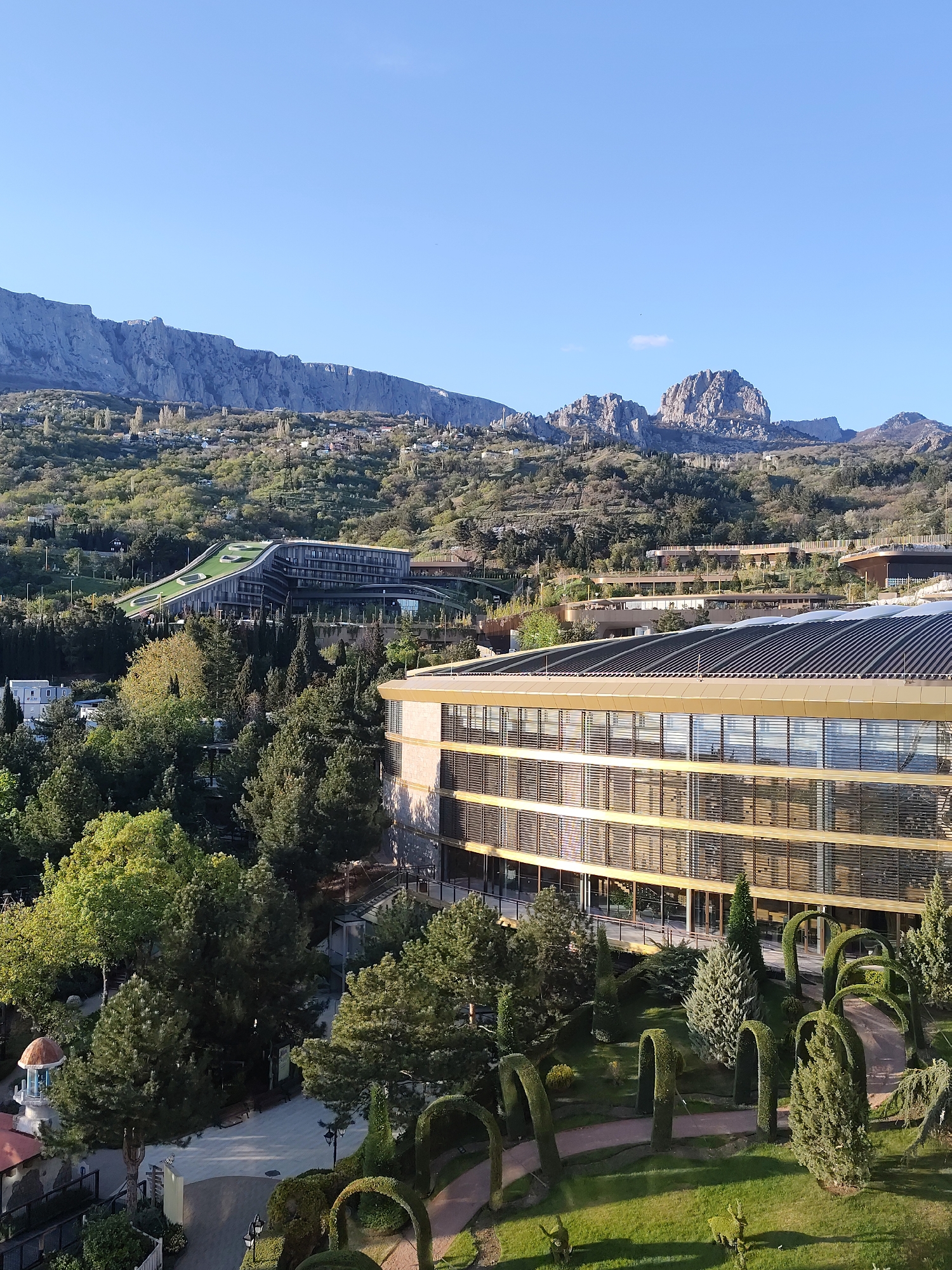 This high-angle, panoramic image captures a scenic landscape featuring modern architecture nestled against a dramatic mountain range under a clear blue sky.
In the background, a rugged, grey mountain range stretches across the horizon, showcasing steep cliffs and prominent peaks. The sky above is bright and clear, indicating fair weather.
The mid-ground is characterized by a verdant hillside densely covered with trees of varying shades of green. Several contemporary buildings are integrated into this natural setting. To the left, a distinctive modern building features a gently sloping green roof, designed to blend with the landscape. Further right and slightly lower, another large, multi-story building, likely a hotel or resort, dominates the foreground. This building boasts a striking gold or bronze-toned facade with numerous windows reflecting the sunlight, and a dark, multi-sectioned roof. Surrounding it is a meticulously landscaped garden with lush green lawns, abundant trees, and intricate topiary art, including arched hedges and shapely shrubs. A paved path winds through this garden. In the lower-left corner, a small, white gazebo with an orange, domed roof is visible.
The scene depicts no discernible activity or people. The clear blue sky and the distinct shadows cast by the buildings and trees suggest it is late morning or early afternoon. The location appears to be a resort or well-developed area within a mountainous region, consistent with the provided context of Симеизский поселковый совет, Ukraine. No visible text is present in the image.
