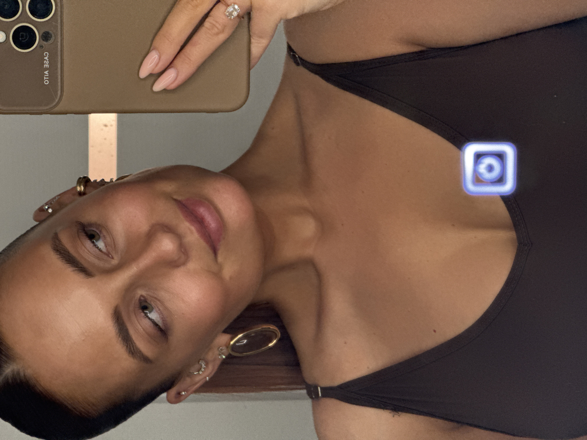 A rotated close-up photograph shows a woman holding a cell phone, presumably taking a selfie. The phone's case is light brown, and her fingernails are manicured with a nude polish. She is wearing a dark brown top. The background appears to be an indoor setting, with a wall visible in the upper left. There is no discernible indication of the city of Chaikovsky, Russia, in the image.